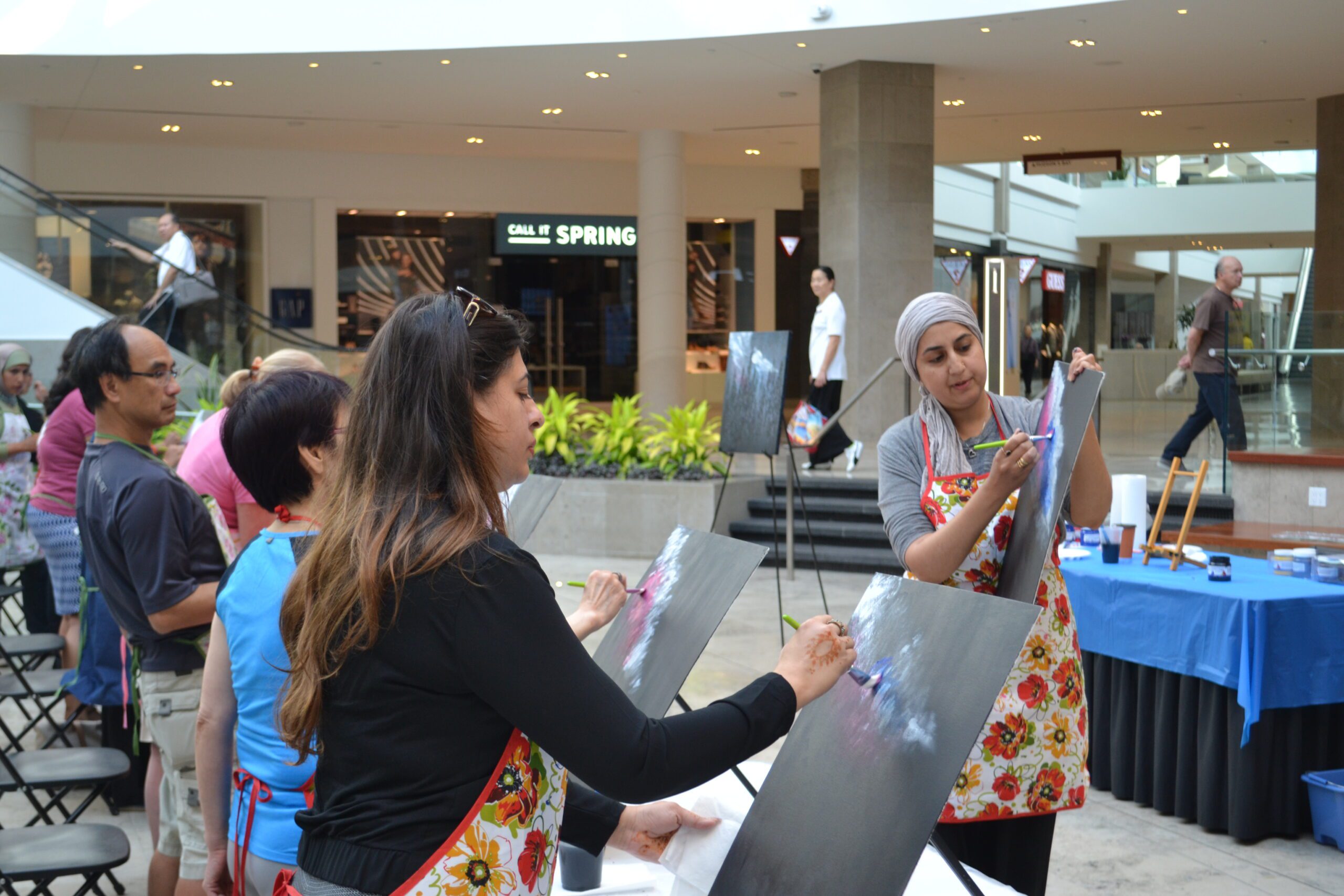 A group of people participating in a public painting workshop inside a bright, modern shopping mall. In the foreground, two women wearing floral aprons are painting colorful abstract designs on black canvases. Other participants stand in line nearby, while shoppers and storefronts are visible in the background.