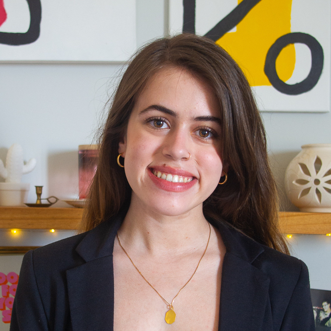 Sabrina Cunha, Multimedia Designer, smiles, wearing a black blazer in front of shelving and wall art.