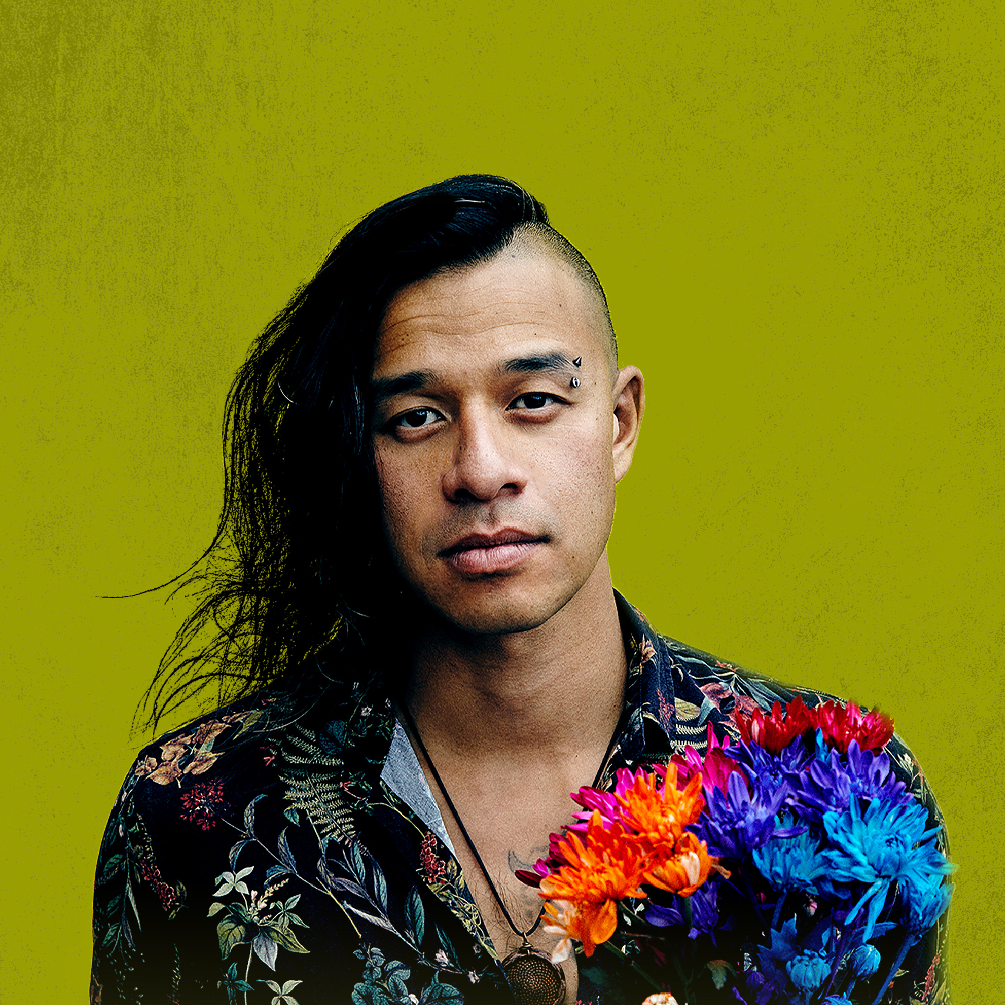 Headshot of Tennyson King. He wears a floral shirt, holding a colourful orange, purple, red, and blue bouquet against a pea green background.