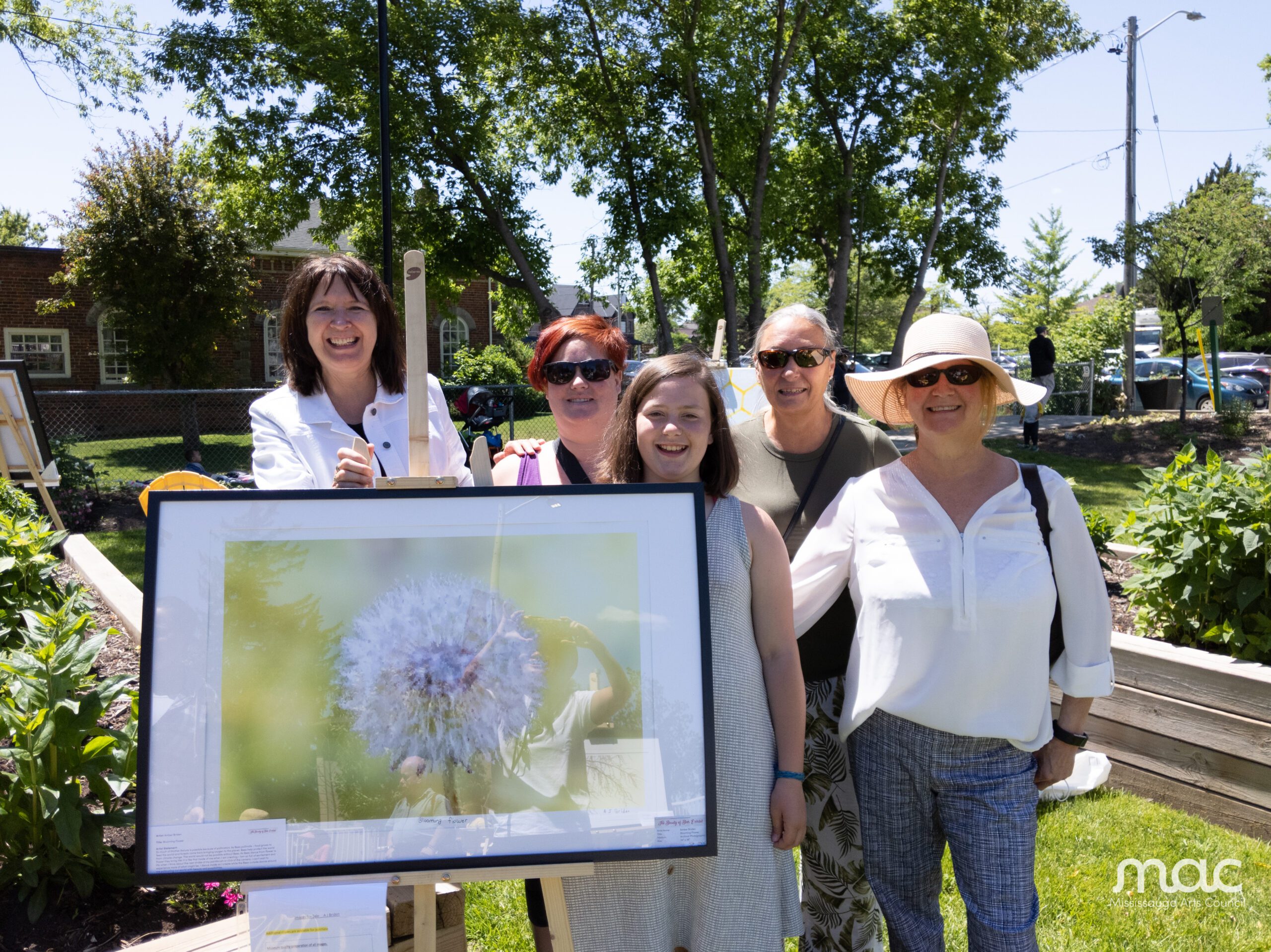 Five women smile together behind a framed photograph of a white dandelion seed head at an outdoor art event in a garden.