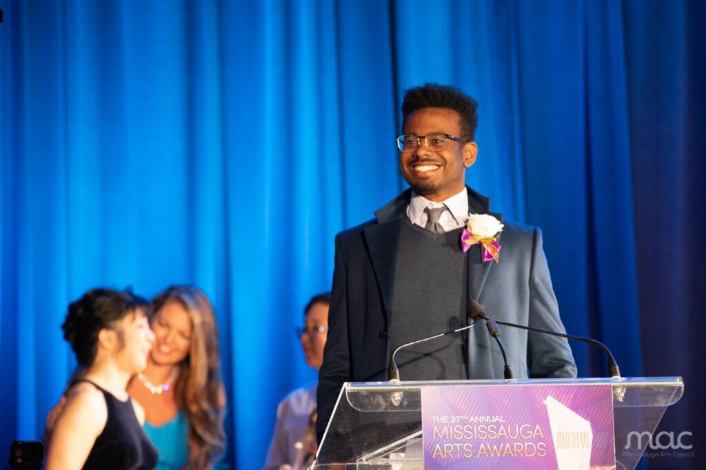 A young man smiles on stage in front of the podium at the 2022 Mississauga Arts Awards.