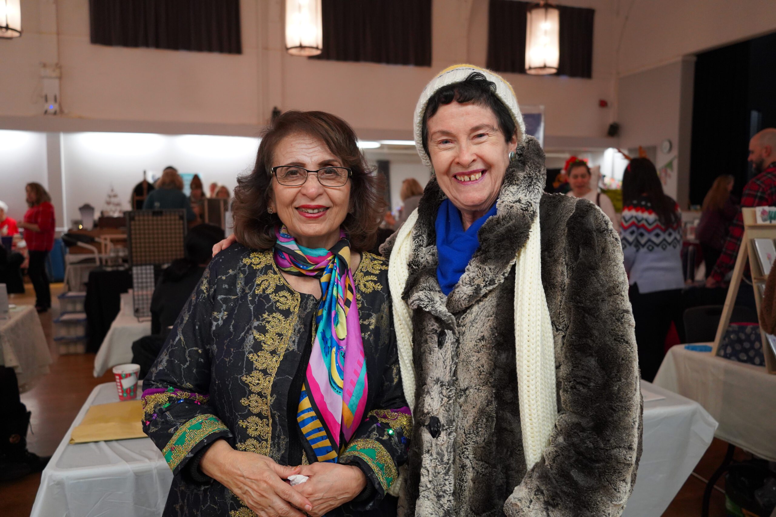 Two women stand close together and smile for a portrait at an indoor market. The woman on the left wears glasses and a black jacket with gold embroidery and a colourful scarf; the woman on the right wears a fuzzy grey coat, a blue scarf, and a white knit hat.