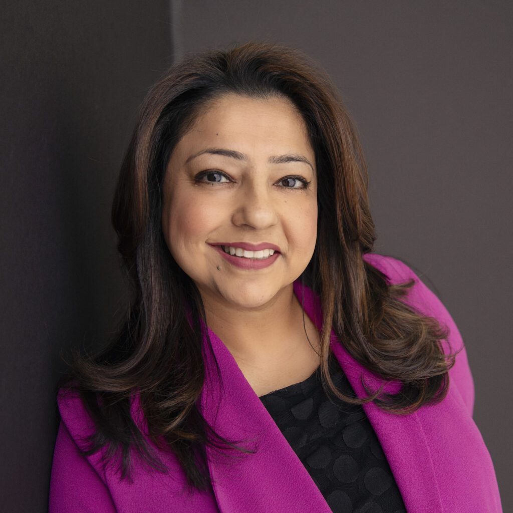 Sumeeta Kohli smiles wearing a bright magenta blazer in front of a dark brown backdrop.