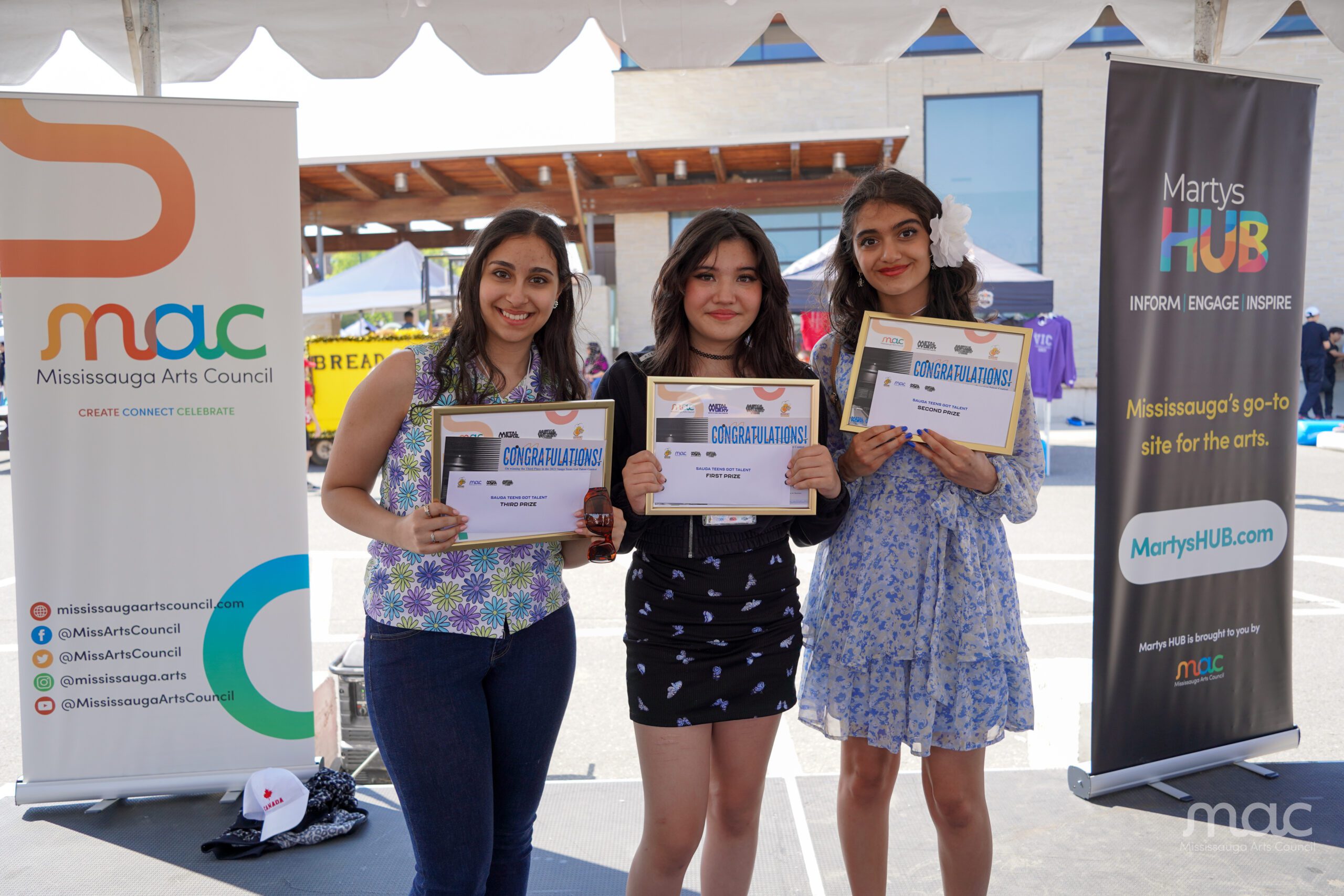 Three smiling young women stand together outdoors, each holding a framed "Congratulations!" certificate. They are positioned between a Mississauga Arts Council (MAC) banner and a Martys HUB banner.