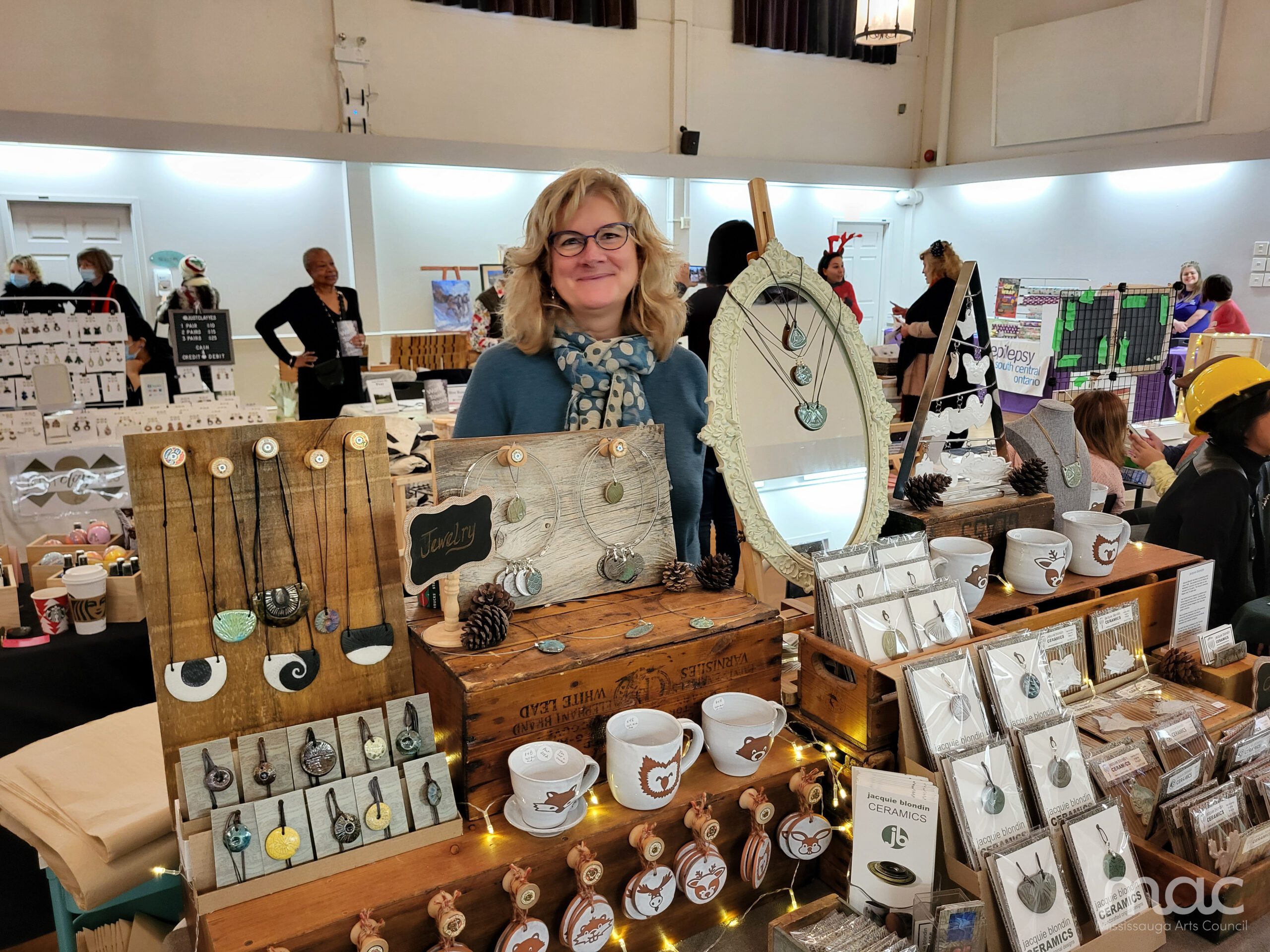 A smiling woman with blonde hair and glasses stands behind a wooden display table at a craft market. She wears a blue sweater and a polka-dot scarf. Her stall features handmade jewelry, ceramic mugs with animal illustrations, and an ornate white oval mirror displaying necklaces. A smiling woman with blonde hair and glasses stands behind a wooden display table at a craft market. She wears a blue sweater and a polka-dot scarf. Her stall features handmade jewelry, ceramic mugs with animal illustrations, and an ornate white oval mirror displaying necklaces.