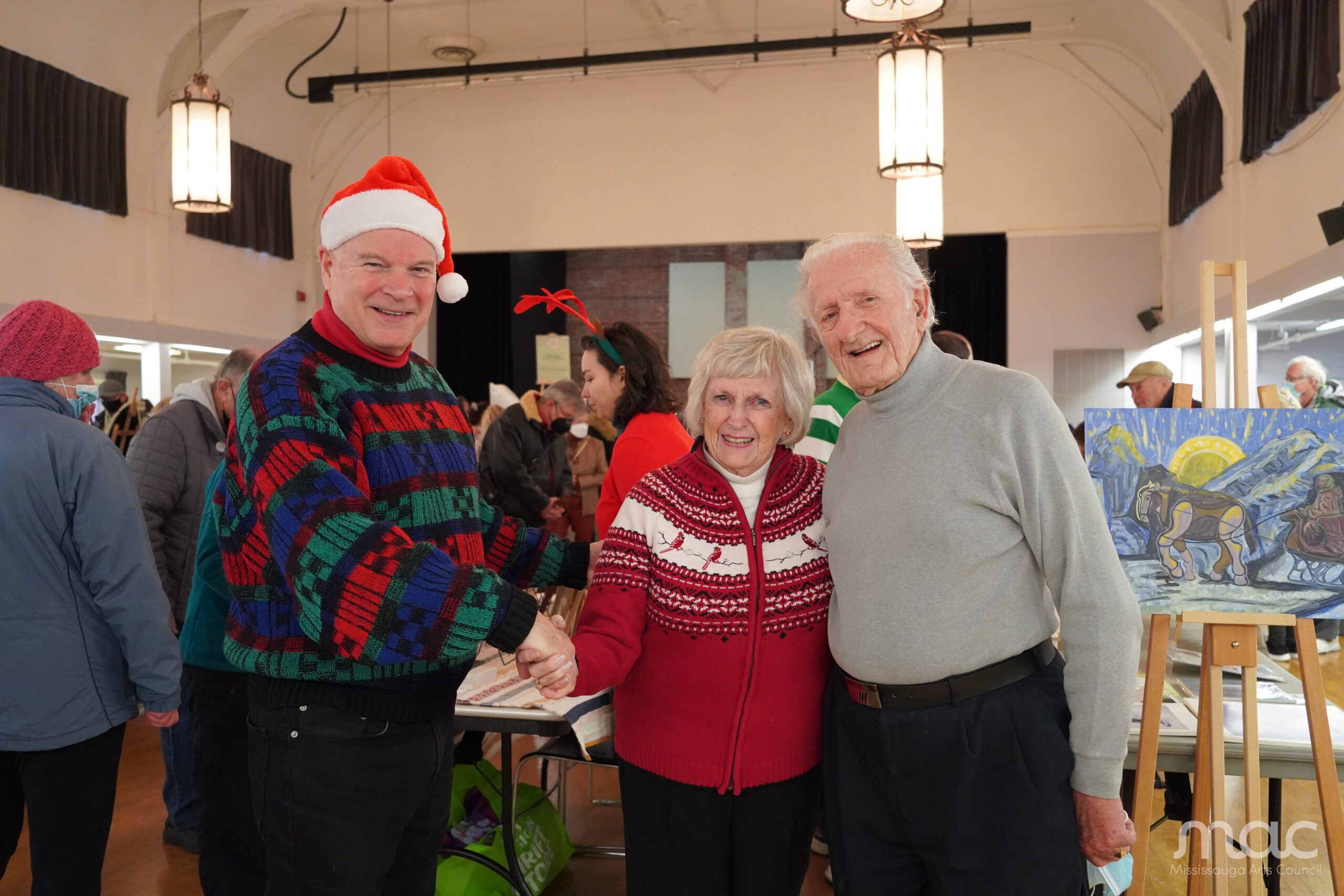 Three cheerful seniors pose together at a Christmas market. A man on the left wears a Santa hat and a festive patterned sweater, shaking hands with a woman in a red cardinal-themed sweater. A second man in a grey turtleneck stands to the right. An easel with a painting is visible in the background. Three cheerful seniors pose together at a Christmas market. A man on the left wears a Santa hat and a festive patterned sweater, shaking hands with a woman in a red cardinal-themed sweater. A second man in a grey turtleneck stands to the right. An easel with a painting is visible in the background.