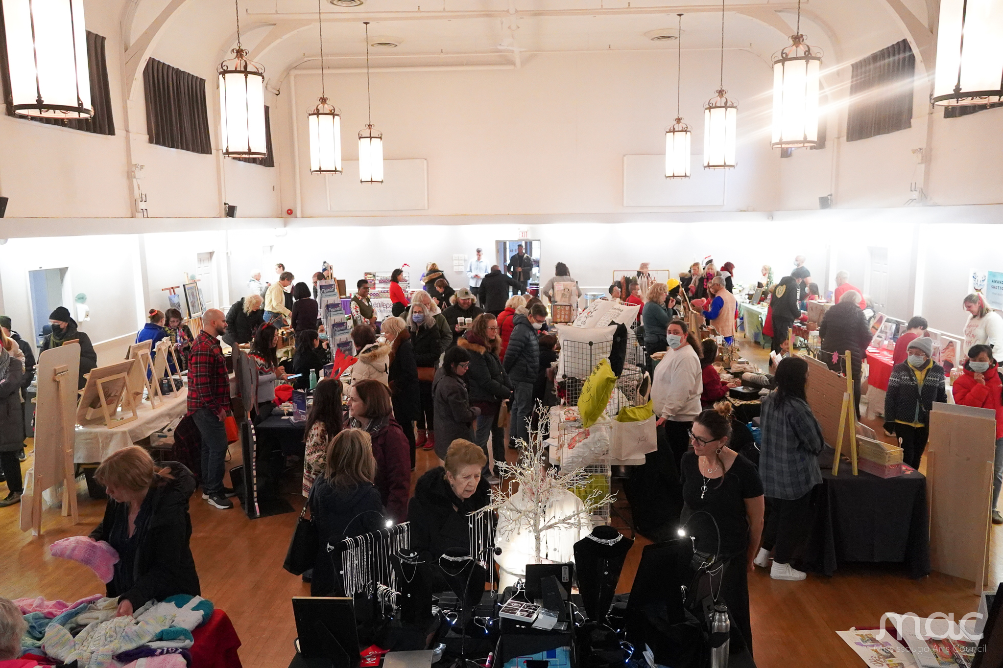A high-angle, wide shot of a bustling indoor holiday market held in a large hall with high ceilings and hanging lantern lights. Numerous vendors have set up tables filled with crafts, and many shoppers are browsing the aisles in winter clothing. A high-angle, wide shot of a bustling indoor holiday market held in a large hall with high ceilings and hanging lantern lights. Numerous vendors have set up tables filled with crafts, and many shoppers are browsing the aisles in winter clothing.