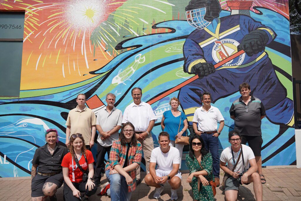 A group of individuals stand and squat smiling in front of a large mural that depicts fireworks, a river scene, and a hockey player wearing a Port Credit Hockey Association jersey.