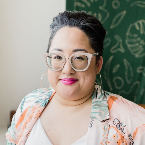Jennifer Rabanillo smiles, wearing a tropical button-up shirt and a white blouse in front of a monstera print 