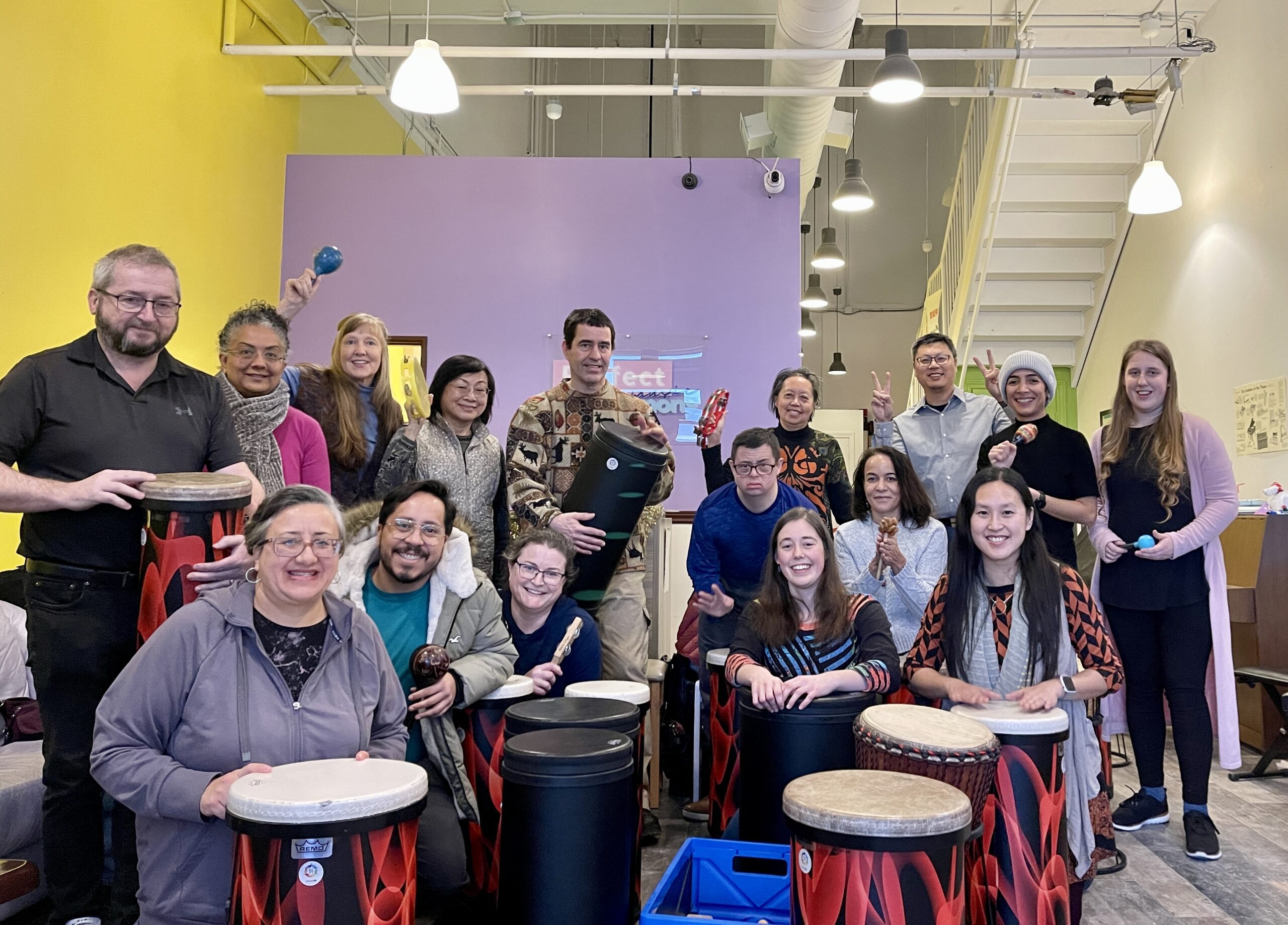 A large, diverse group of people of various ages and abilities sit and stand in a brightly coloured room (yellow and purple walls), posing with various percussion instruments and colourful floor drums.