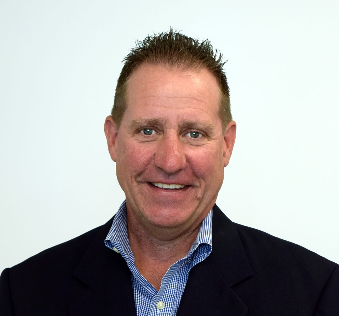 Brian Dec smiles, wearing a black blazer and blue button-up shirt in front of an off-white backdrop.