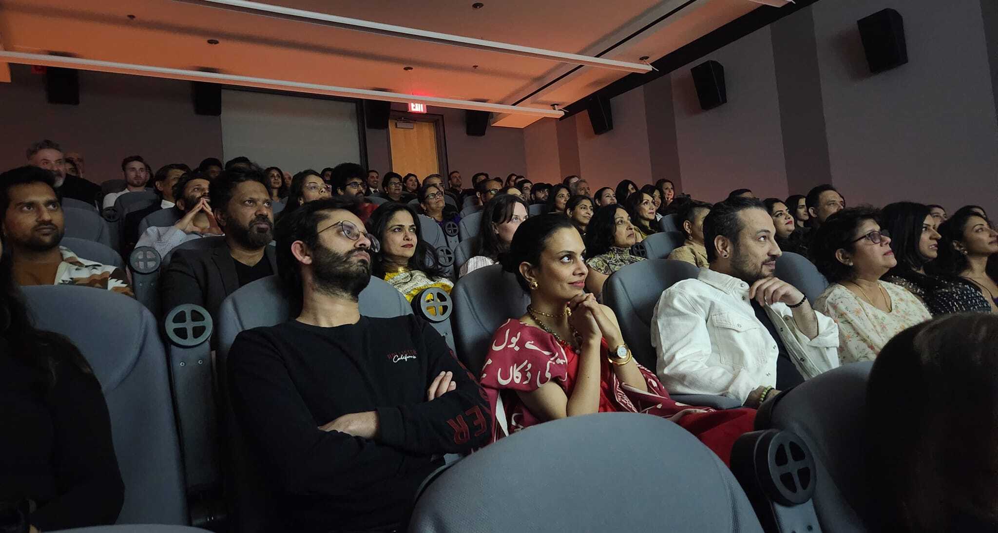 A large, diverse audience sitting in a dimly lit theatre, focused on a screen or performance off-camera.