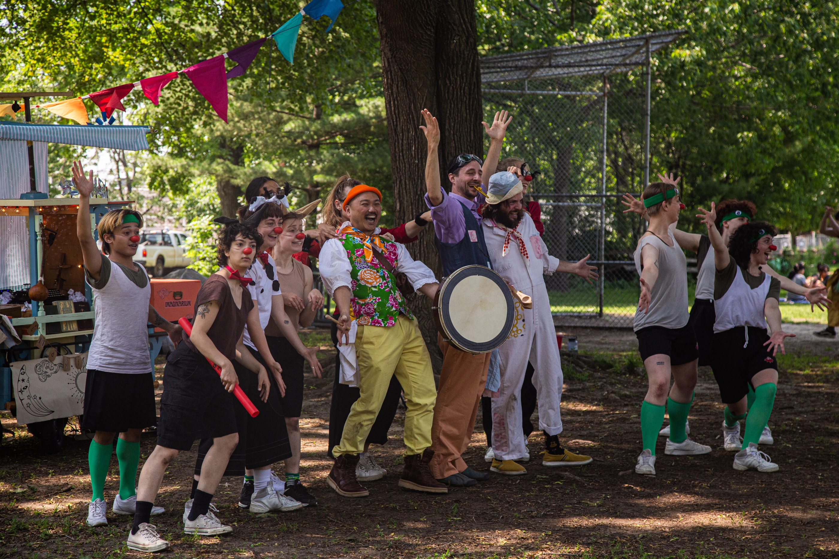 A troupe of performers dressed as clowns with red noses and eclectic costumes dance and cheer in a wooded park area. Some hold drums and props, creating a festive atmosphere.