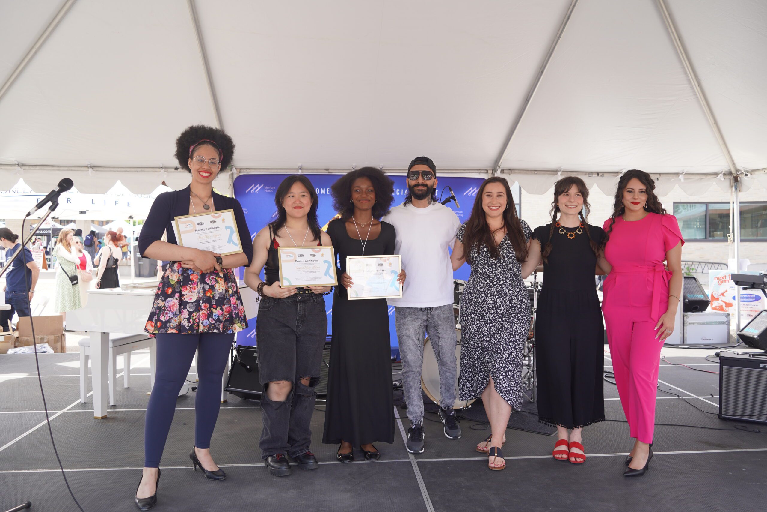 Seven diverse performers stand in a row on an outdoor stage under a white tent. Three of the individuals hold framed award certificates. A white piano and musical equipment are visible on stage behind them.