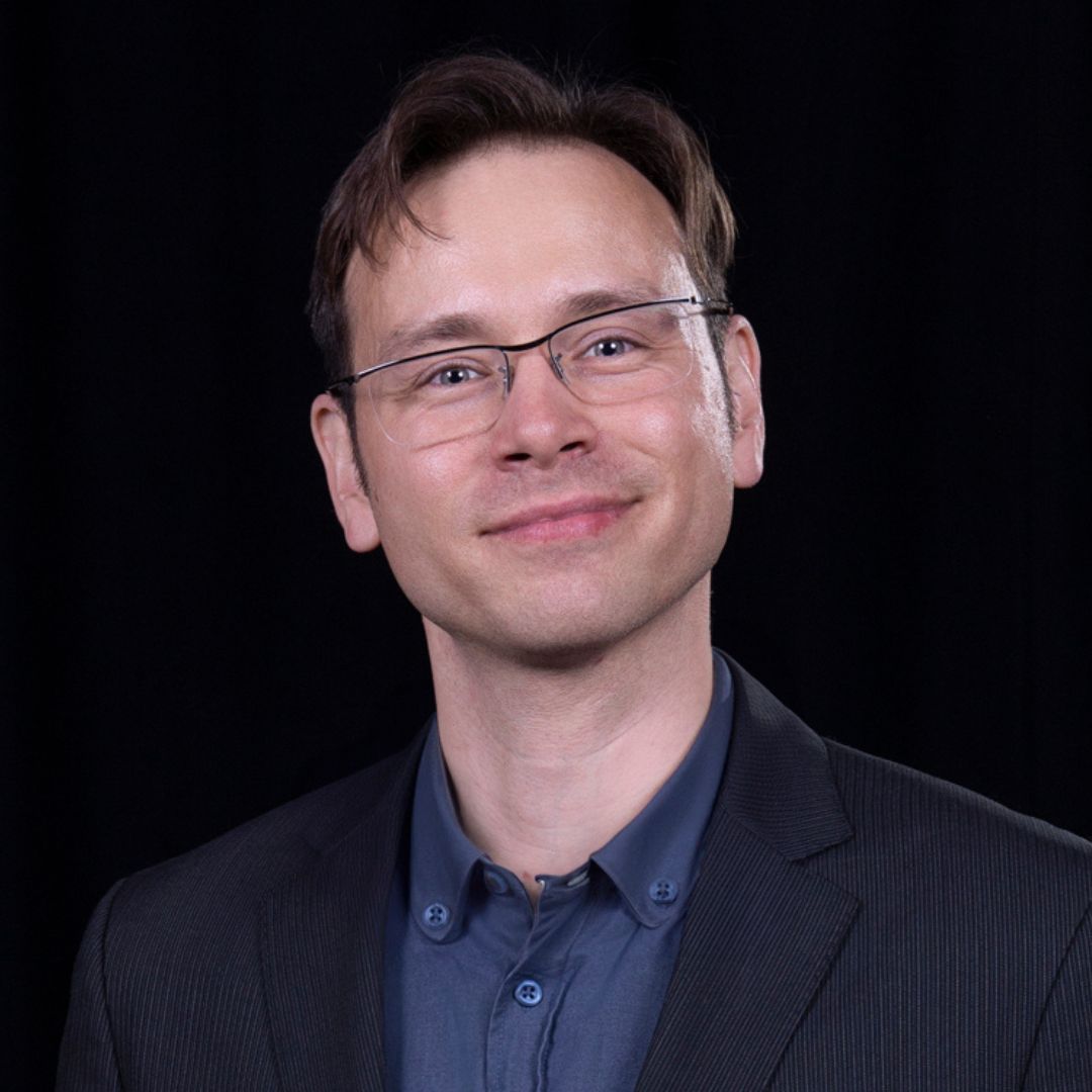 Filip Plaskota smiles, wearing a navy blue blazer over a blue button-up in front of a black backdrop.