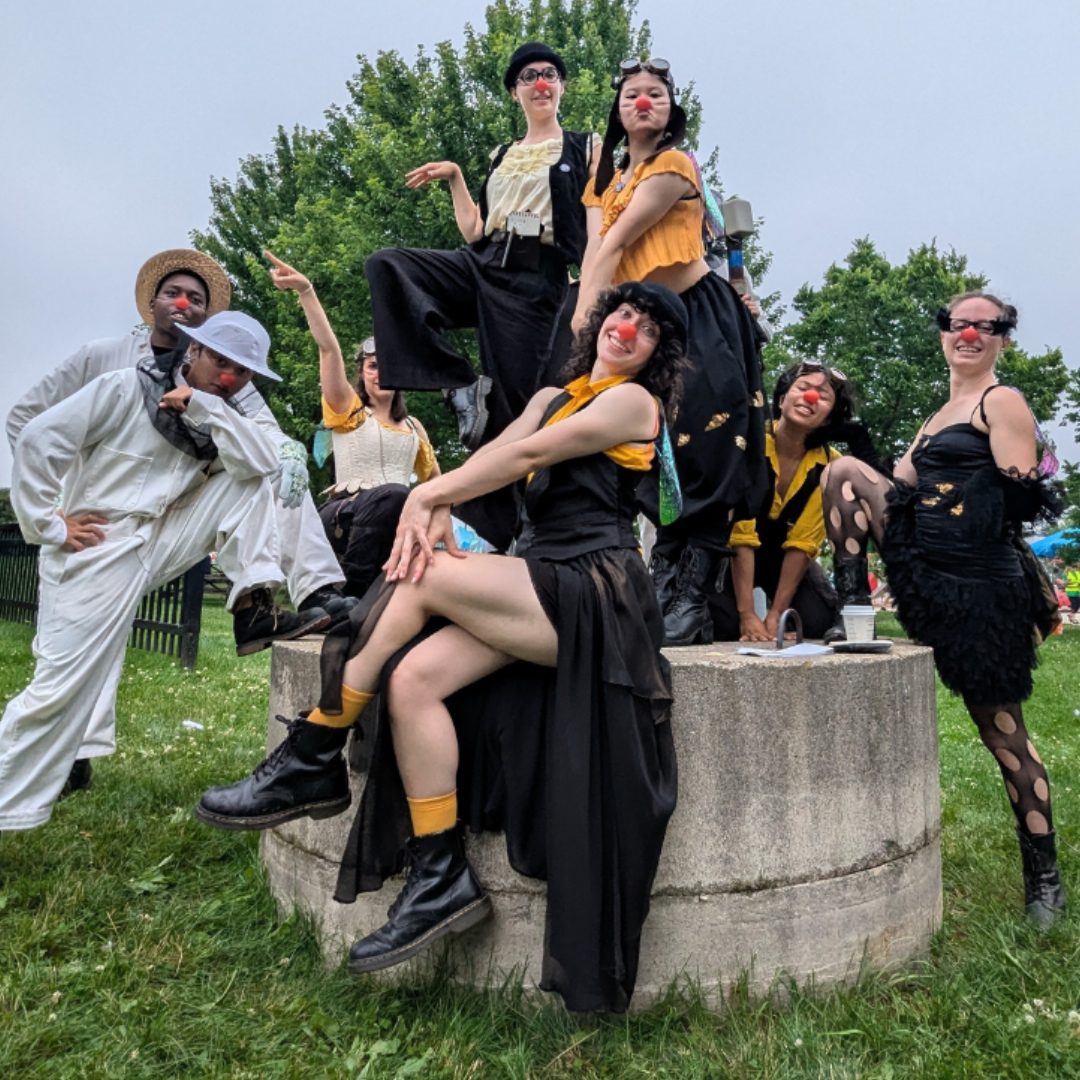 Group photo of dancers dressed as clowns in black and yellow and beekeeping suits. They pose on a concrete well at a public park.