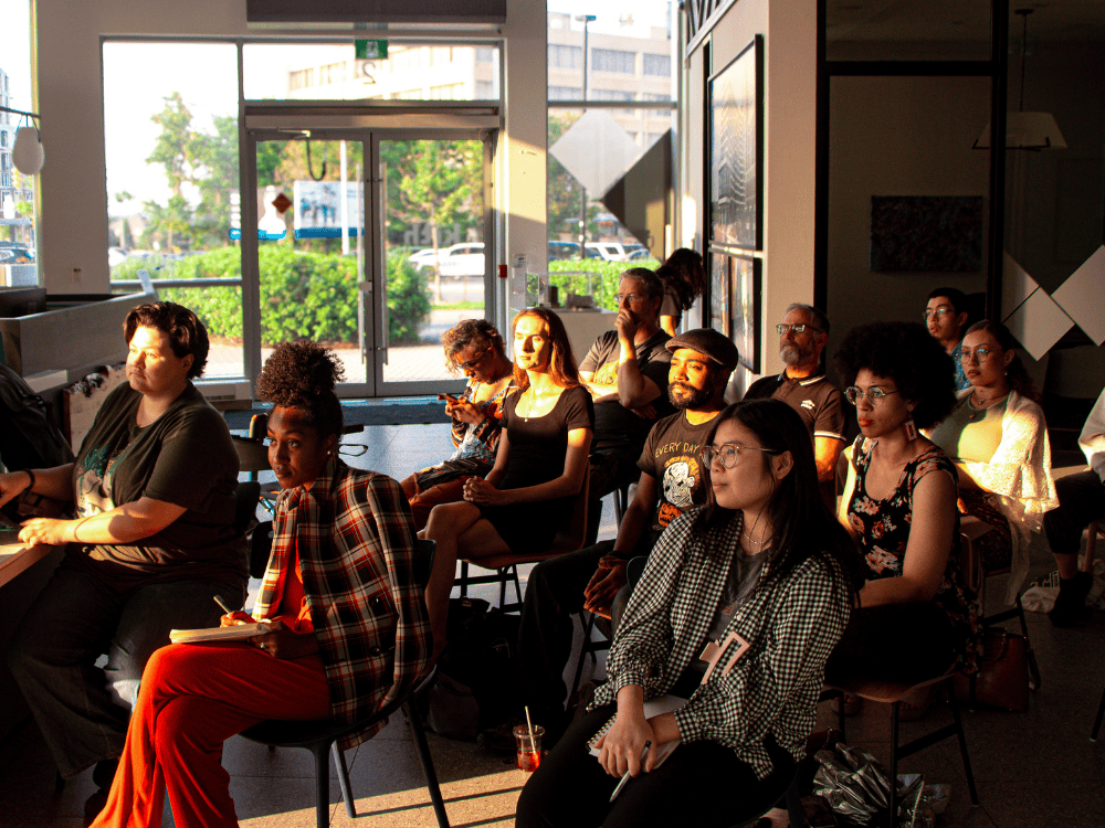 A diverse group of people attentively listening at an Exprose event, sitting in a modern, sunlit space with large windows and an urban backdrop. Some attendees are taking notes while others are focused on the presentation.