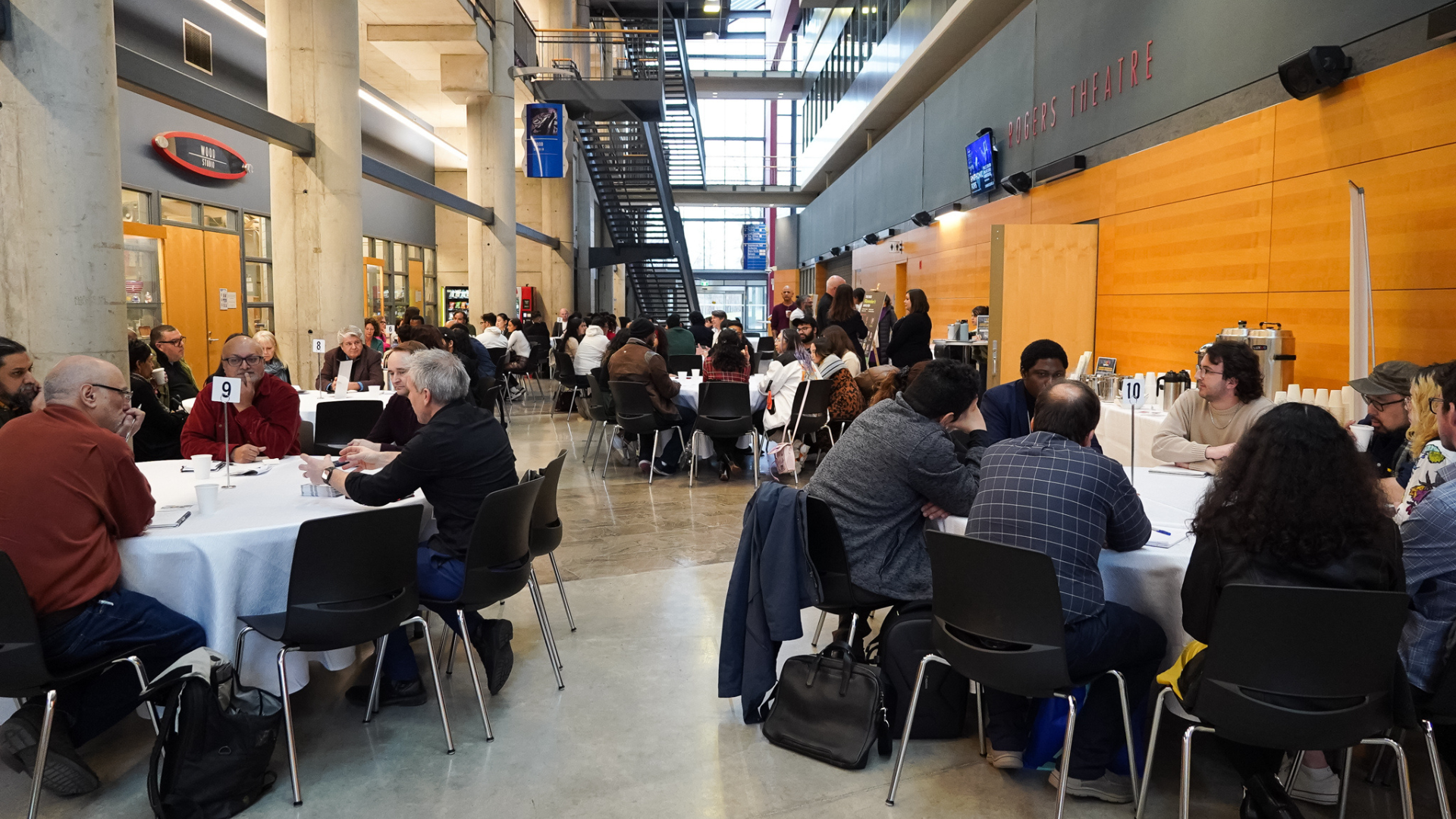 A high-angle shot of a bright, modern atrium with concrete pillars. People are seated at several white round tables for a networking event. A "ROGERS THEATRE" sign is visible on the wood-panelled wall in the background. A high-angle shot of a bright, modern atrium with concrete pillars. People are seated at several white round tables for a networking event. A "ROGERS THEATRE" sign is visible on the wood-panelled wall in the background.