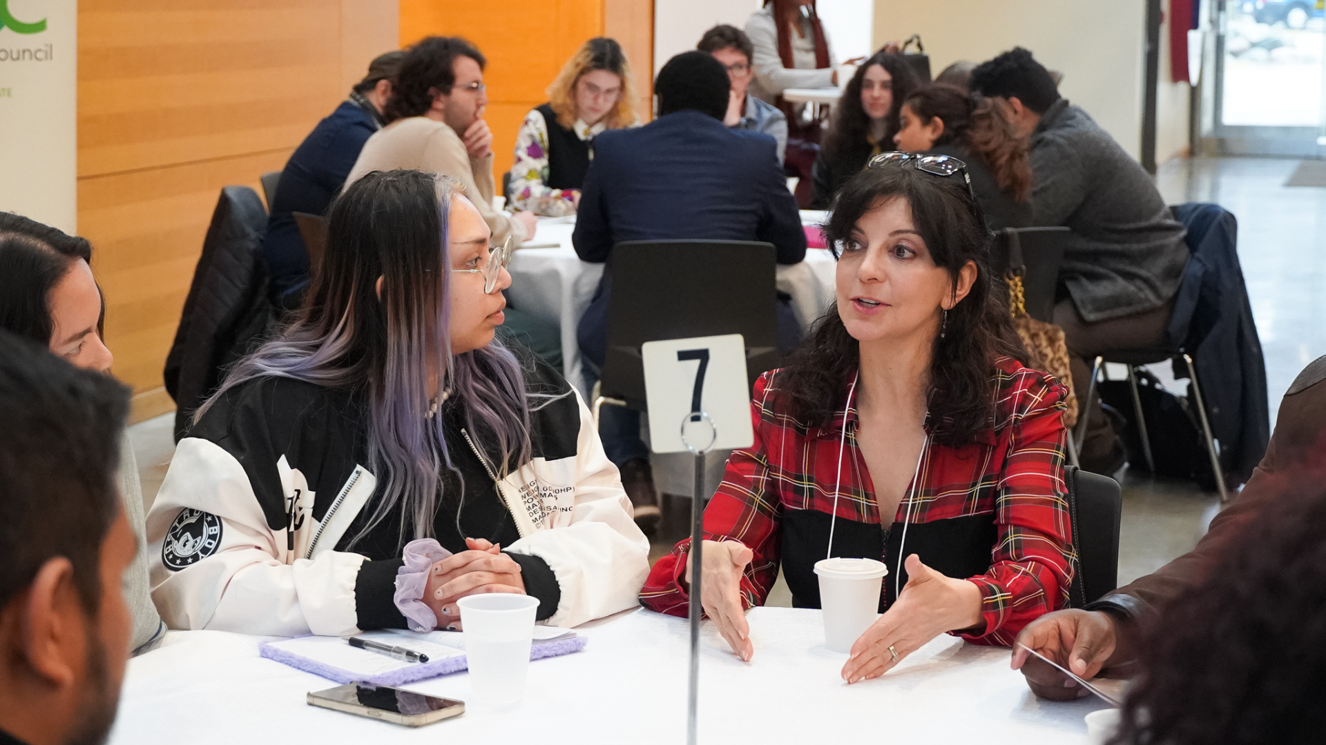 A woman in a red plaid shirt gestures with both hands while speaking to a younger woman with purple-tinted hair at a roundtable. Other attendees are blurred in the background of the bright hall. A woman in a red plaid shirt gestures with both hands while speaking to a younger woman with purple-tinted hair at a roundtable. Other attendees are blurred in the background of the bright hall.