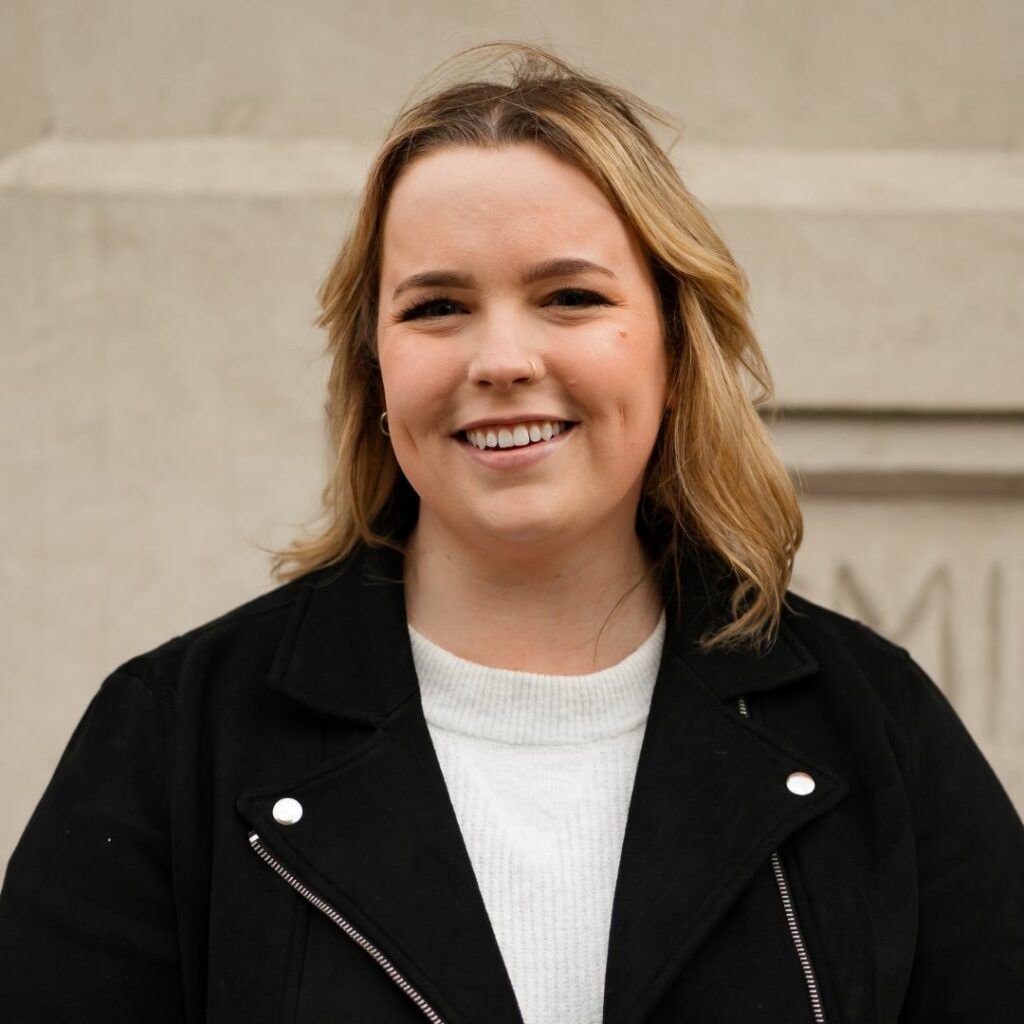 Sherri Parliament, Communications Manager and Music Strategist, smiling wearing a black jacket and white top, standing in front of a cement building.