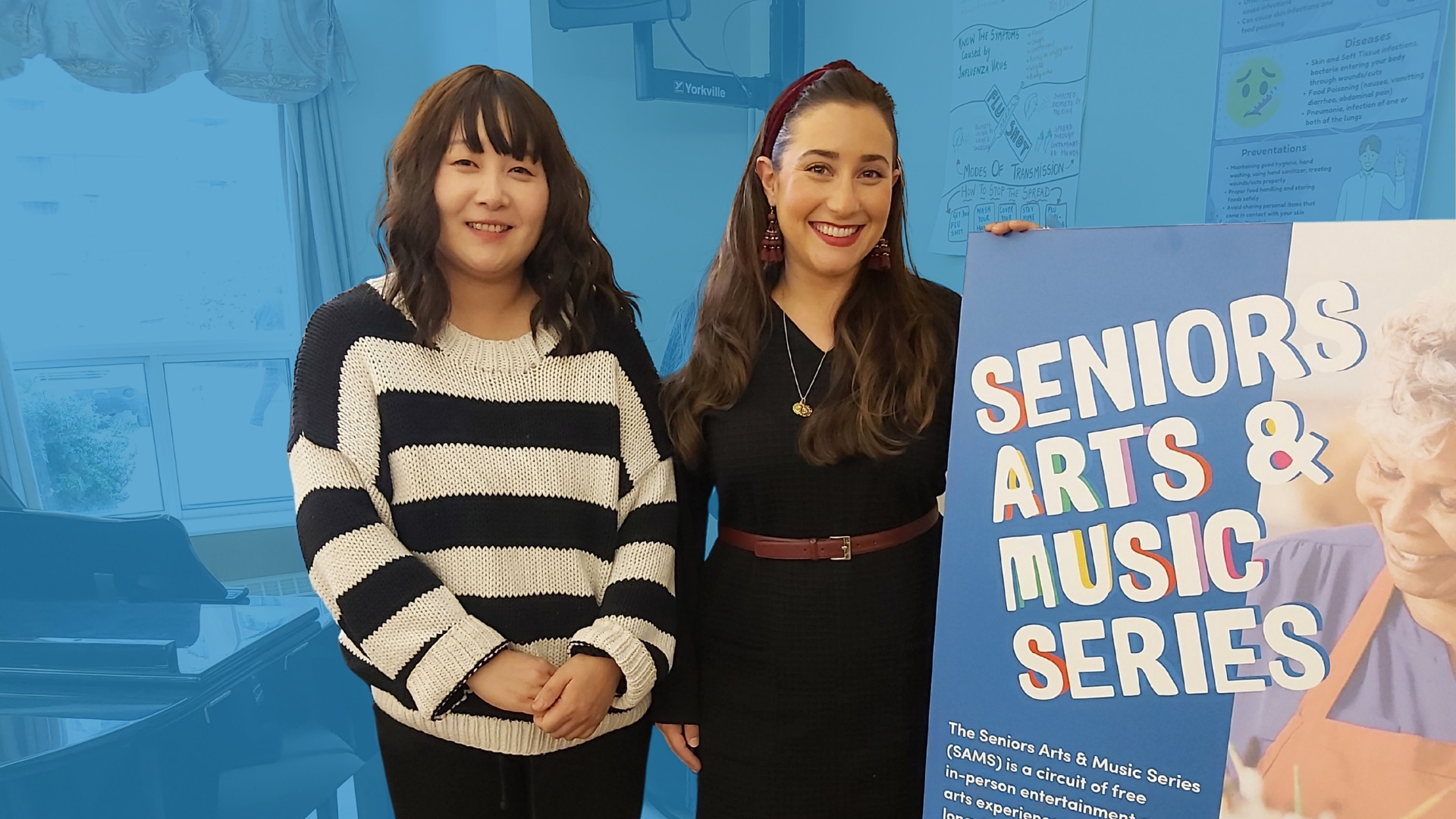 Two women stand beside a piano in a long-term care community room. The woman on the right holds a poster that reads "Seniors Arts & Music Series"