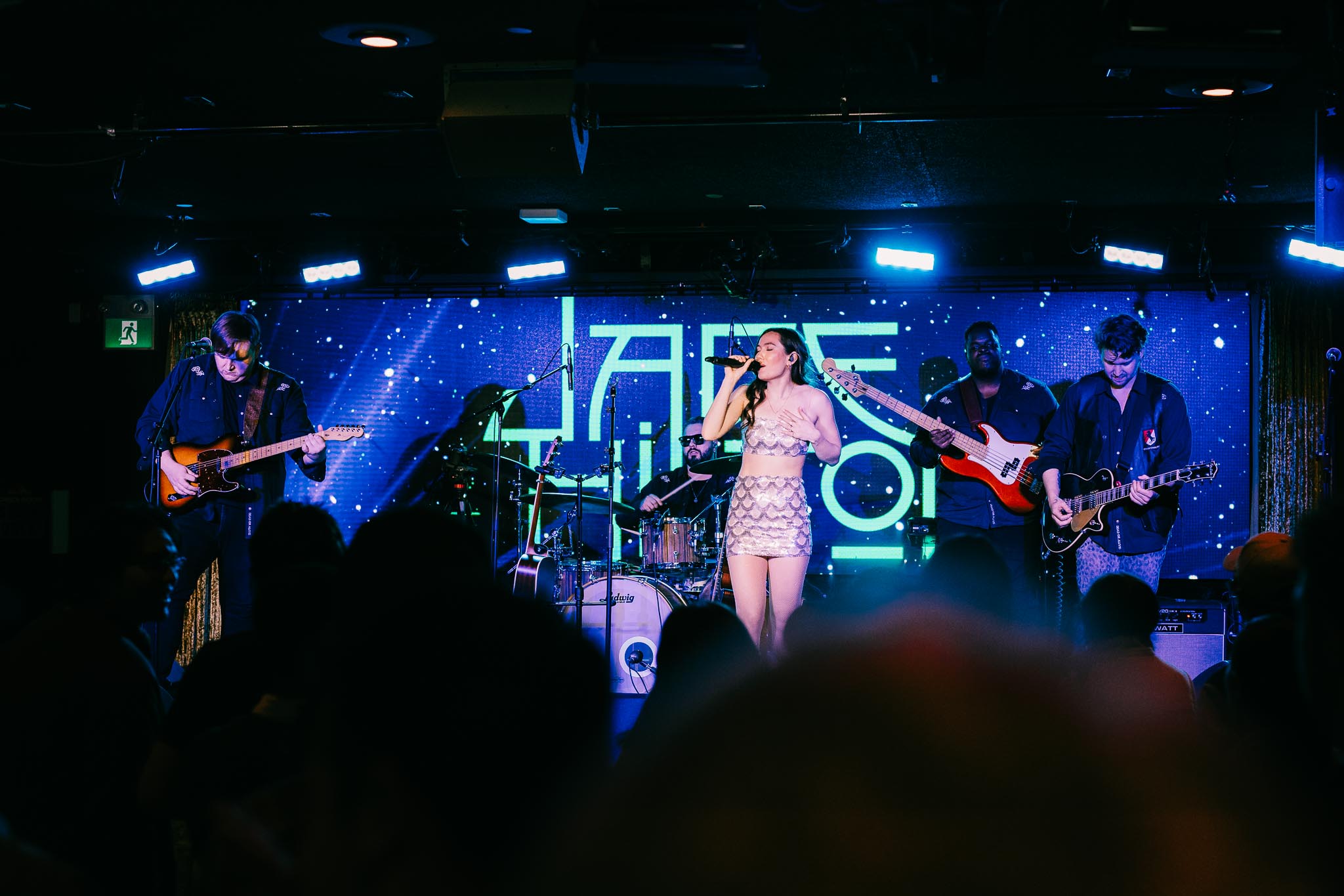A woman singer in a sparkling two-piece outfit performs on a dark stage with a full band. A large digital screen behind them displays "Jade Hilton" in glowing neon letters against a starry blue background.