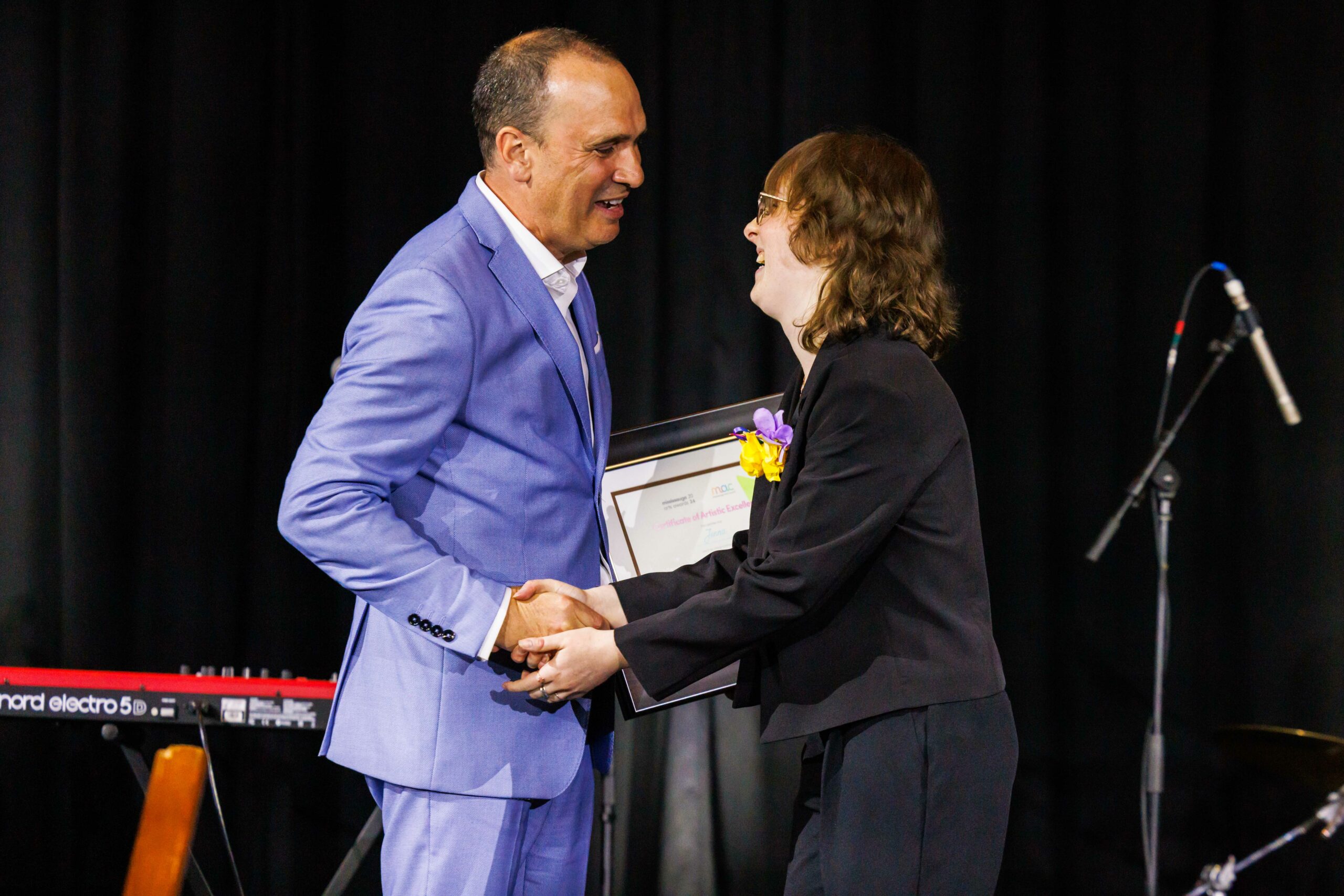 A joyful moment at the Mississauga Arts Awards where a man in a light blue suit presents a framed certificate of excellence to a smiling award recipient. Both are shaking hands on a stage equipped with musical instruments, set against a professional black backdrop.