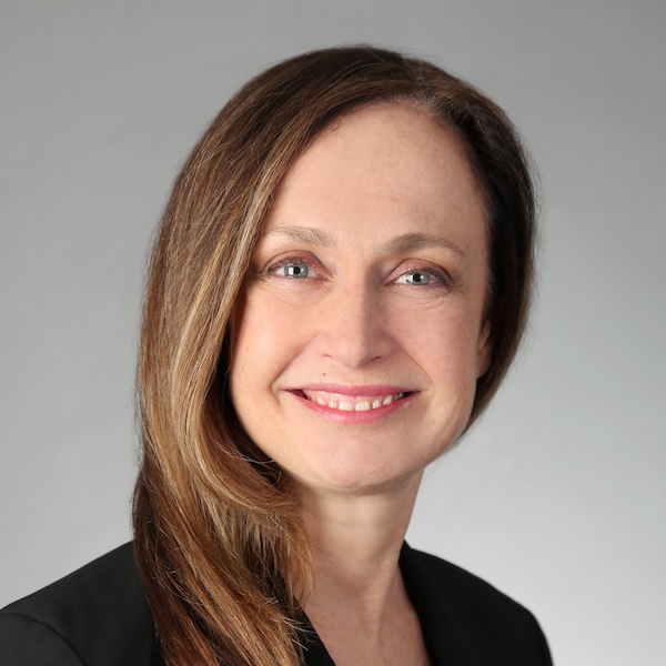 Janey Obad smiles in a black blazer in front of a grey backdrop.