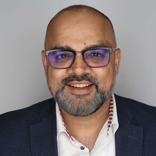 Usman Khan smiles, wearing a blue blazer in front of a grey backdrop.