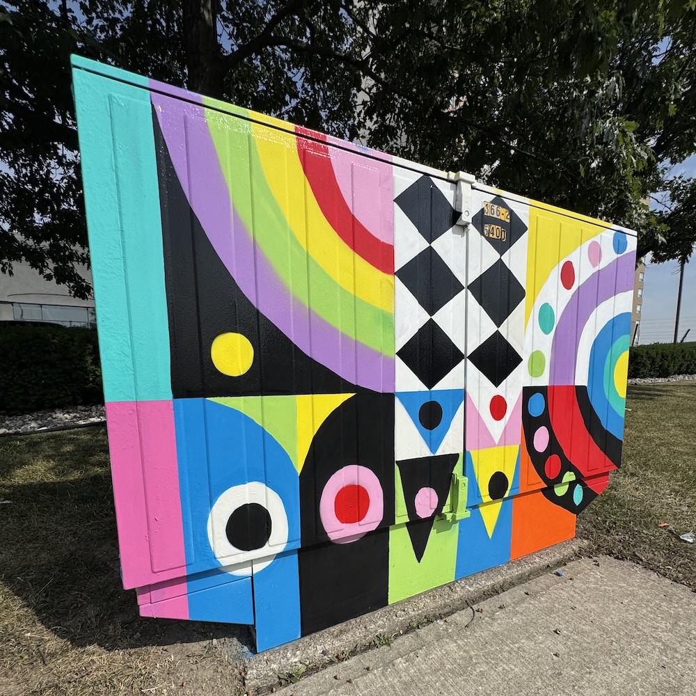 A utility box hand-painted with solid blocks of colour in a geometric pattern. Colours include blue, teal, pink, purple, green, red, orange, black and white.