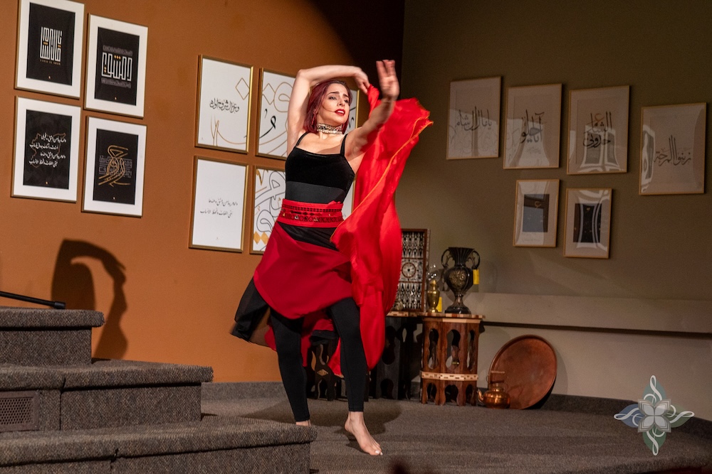 A barefoot dancer in a red and black costume performs a dynamic movement with flowing red fabric in a warmly lit room. Behind her, framed Arabic calligraphy artwork hangs on tan and olive-green walls. Traditional Middle Eastern decor items, including instruments and vases, are visible near the stage.