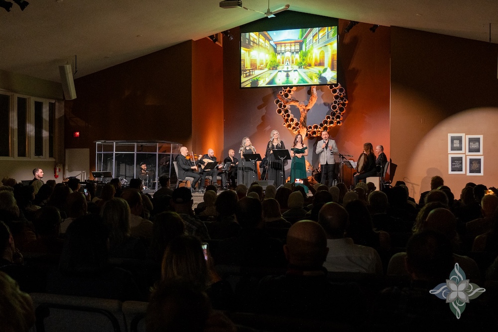 A large audience watches a live performance in a warmly lit venue. A group of singers and musicians is on stage, accompanied by string instruments. A tree-like wall sculpture and a vibrant screen projection showing a courtyard scene provide visual backdrop.