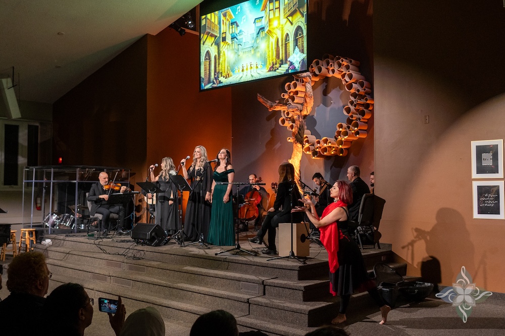 A group of vocalists and instrumentalists perform on stage under a screen displaying an animated Middle Eastern street scene. A dancer in red and black moves expressively on the steps beside them. A sculptural tree and framed Arabic artwork decorate the background.