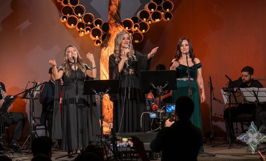 A group of vocalists and instrumentalists perform on stage under a screen displaying an animated Middle Eastern street scene. A sculptural tree decorates the background.