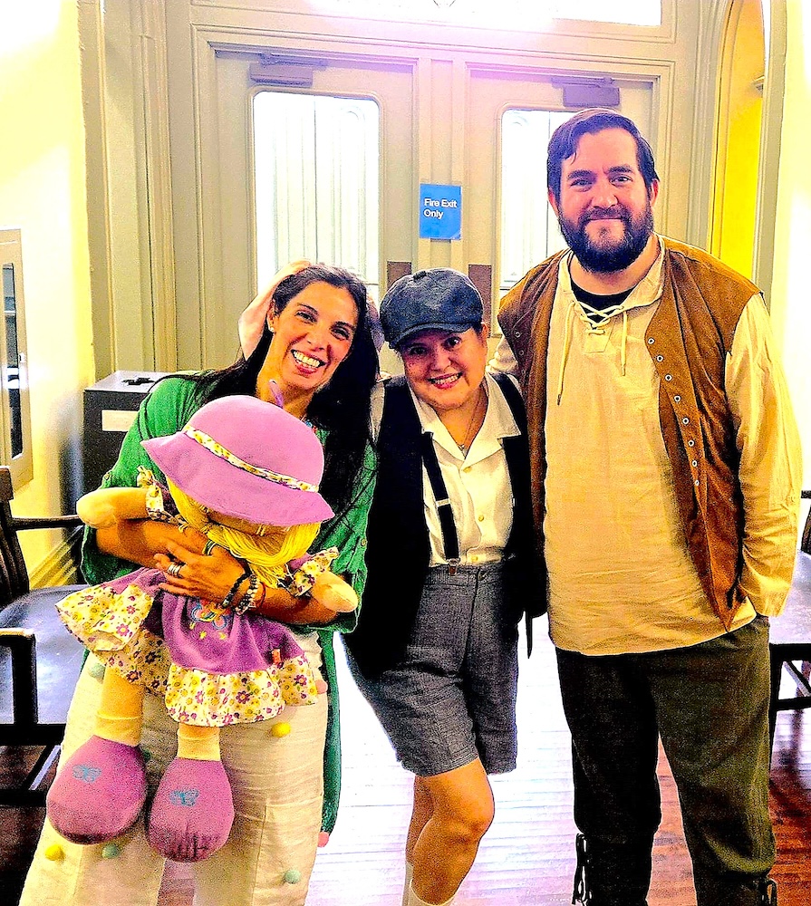 Three actors, dressed in costume and holding props: a doll, smile in front of a makeshift stage.