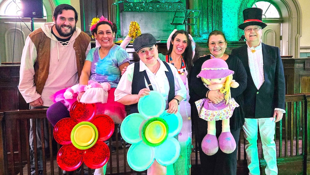 Six actors, dressed in costume and holding props: giant flowers and a doll, smile in front of a makeshift stage.