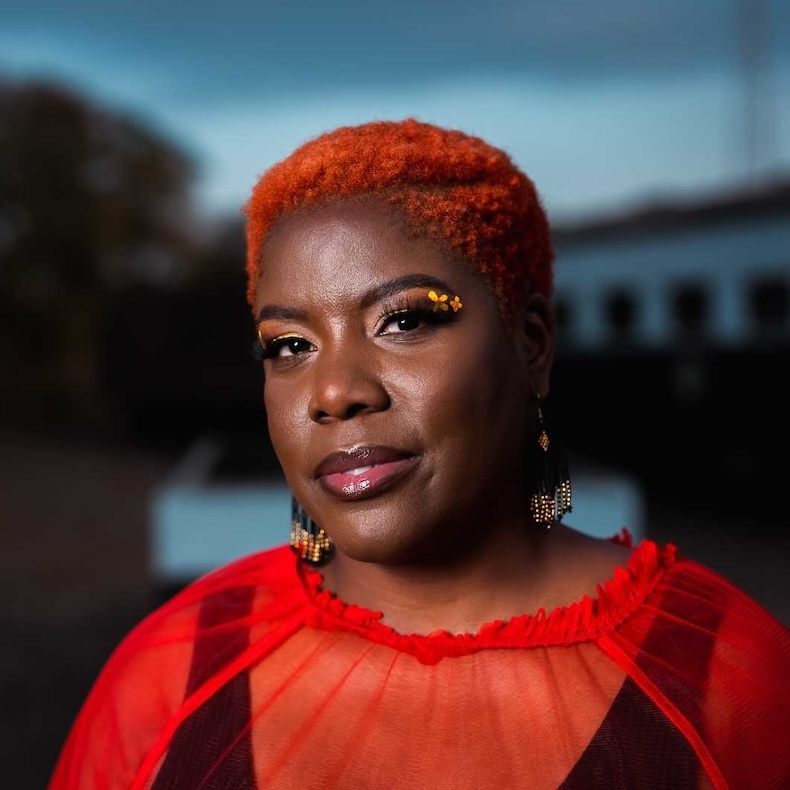 Glodeane Brown, Community Outreach & Production Manager, wearing a red mesh blouse and black tank top in front of a grey/blue outdoor background. 