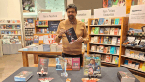A man stands in a bookstore in front of a table with books, promotional items, and an award. He holds a book titled “Shards of Verse” by Pratap Reddy.
