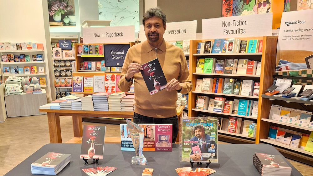 A man stands in a bookstore in front of a table with books, promotional items, and an award. He holds a book titled “Shards of Verse” by Pratap Reddy.