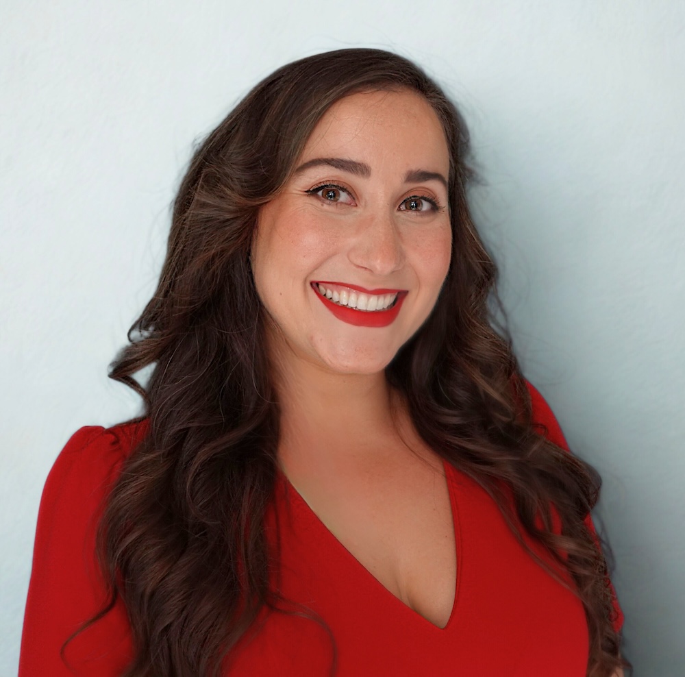 A headshot of Cat Bernardi, who smiles at the camera, wearing a red blouse and matching red lipstick against a white background.