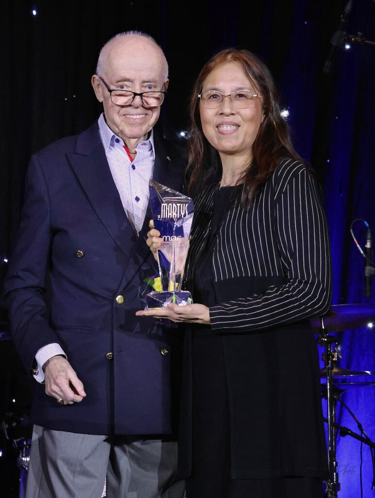 Two people stand on stage at the 2025 MARTY Awards. A woman holds a MARTY trophy.