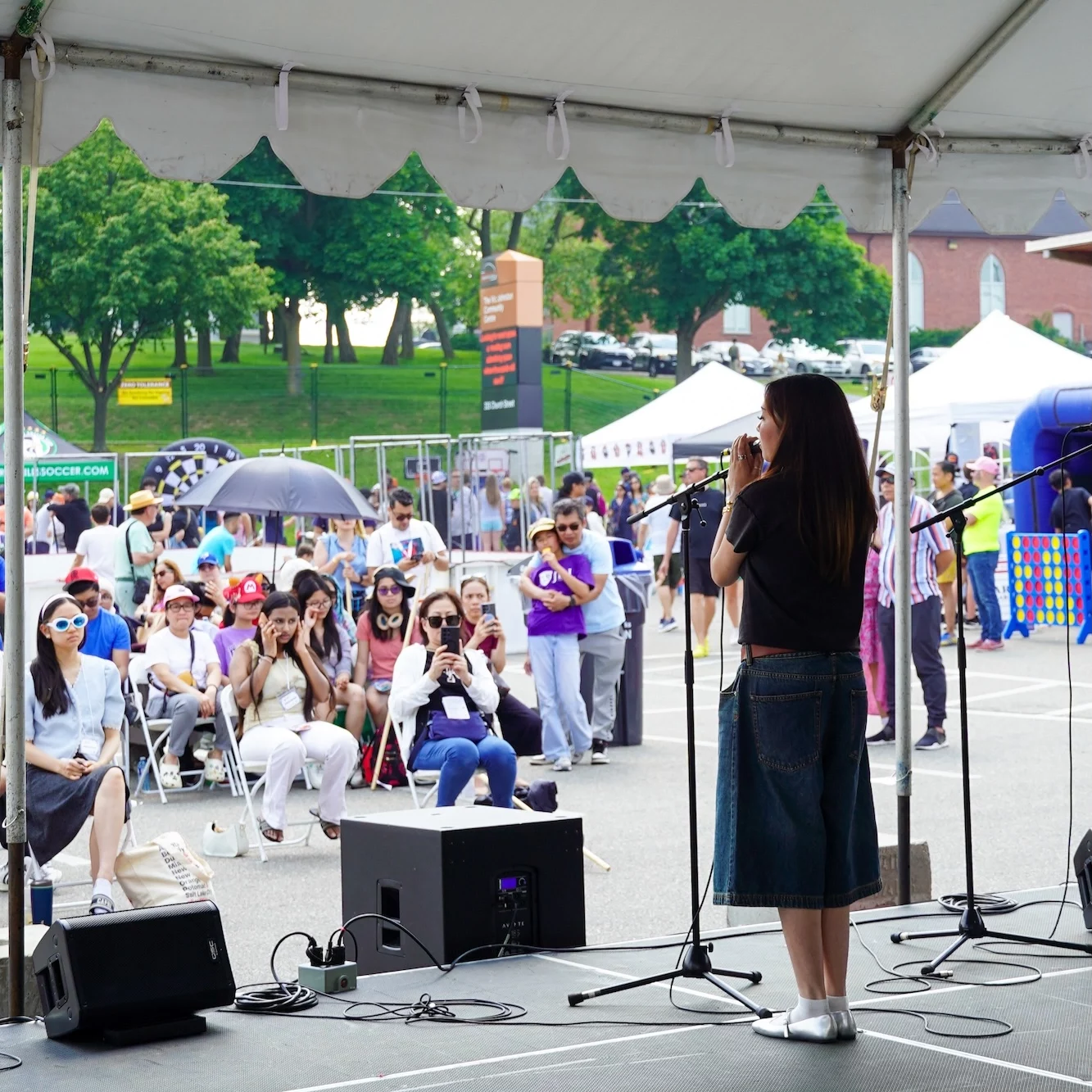 A view from behind a performer on stage looking out at a seated audience watching an outdoor community event.