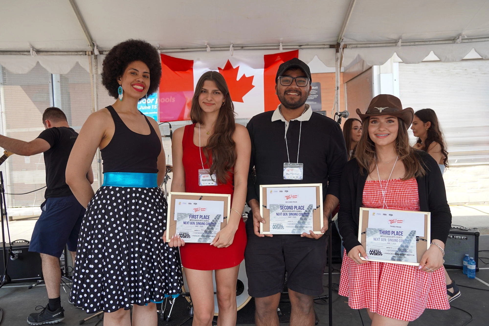 Four people standing together on an outdoor stage; three hold award certificates for first, second, and third place in a singing contest.