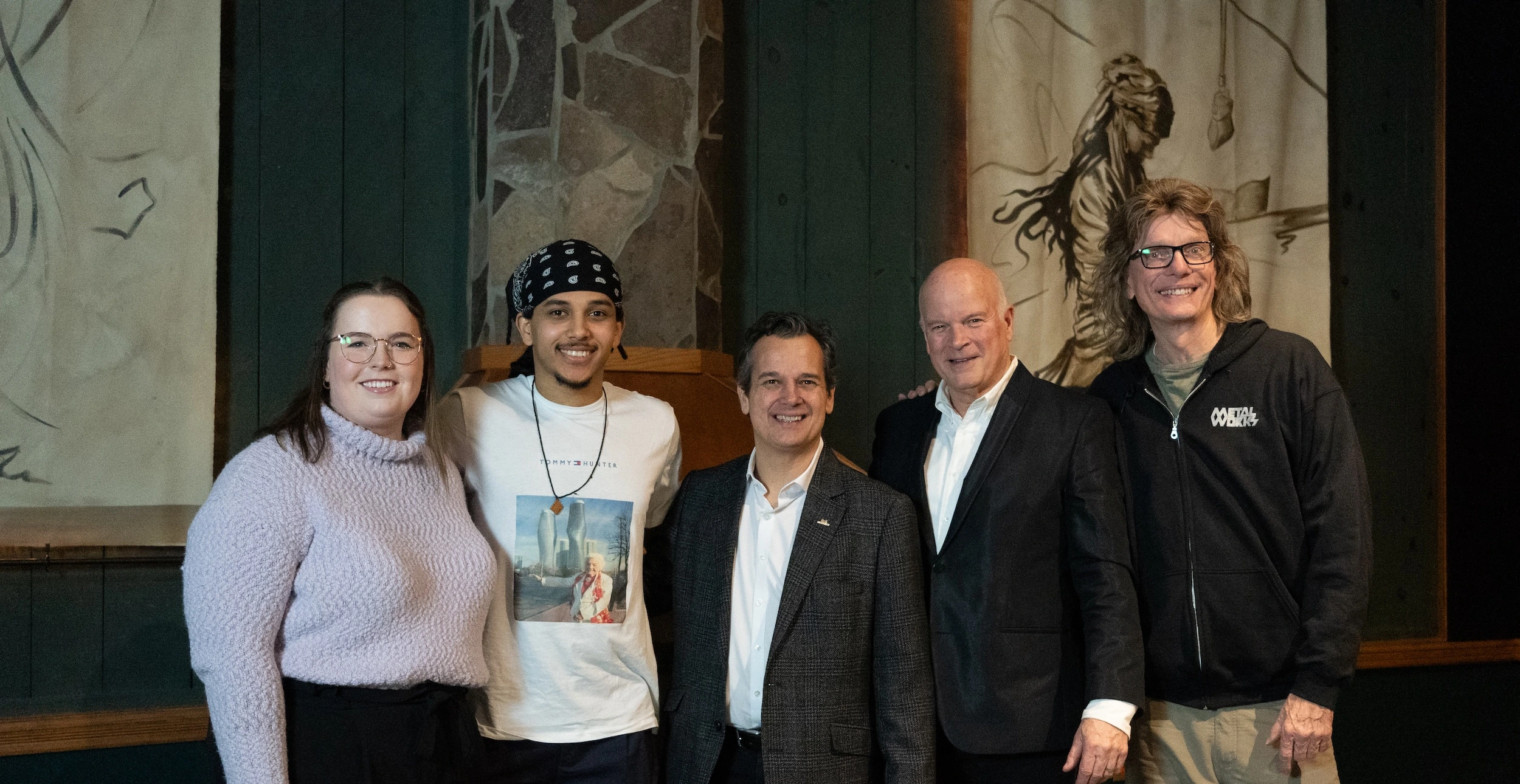 Five people stand together for a group photo in a room with green wooden paneling. The group includes a young woman in a lilac sweater, a young man wearing a black bandana, and three older men in professional attire, the last in a black "Metalworks" hoodie. Large sketch-style artworks hang on the walls behind them.