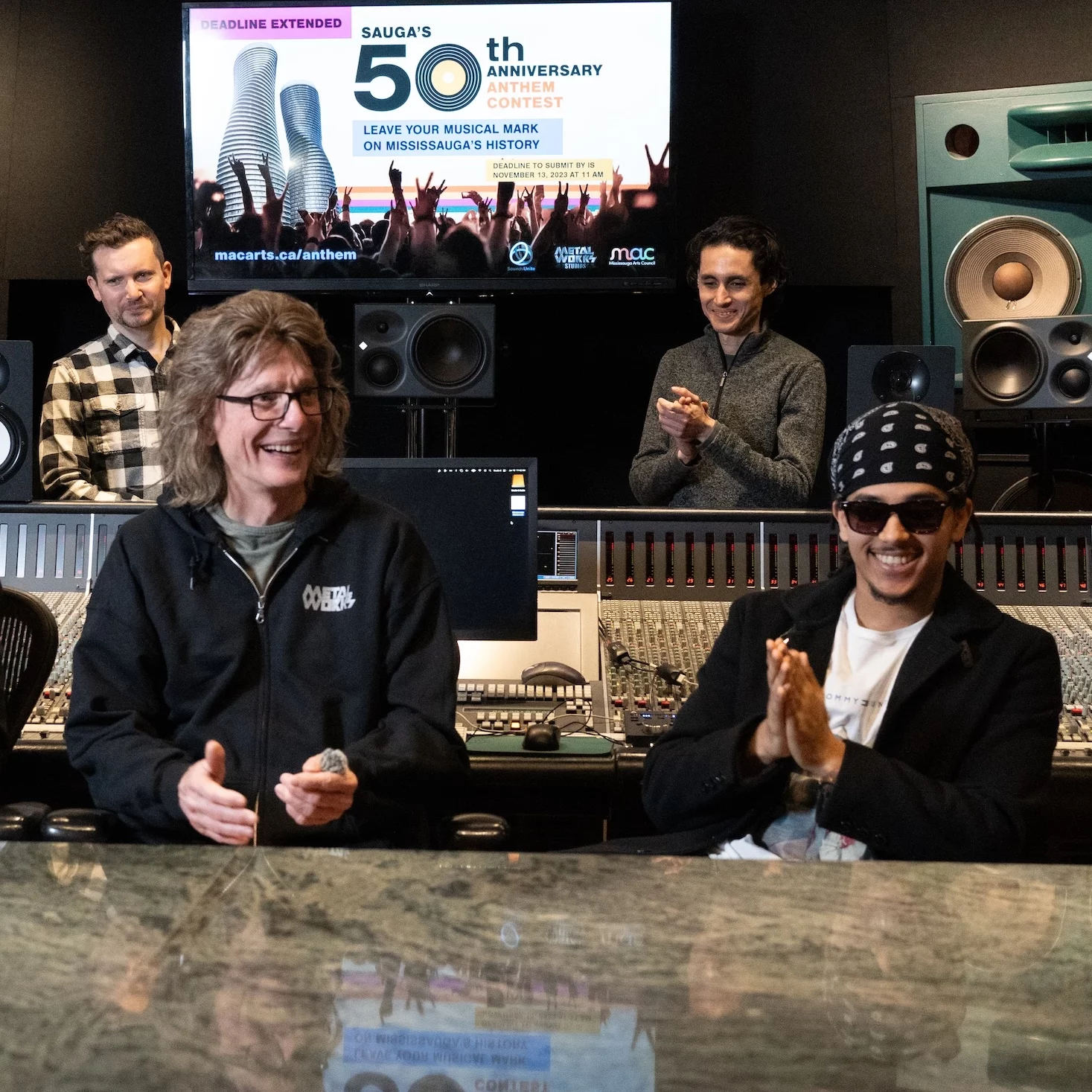 Three men sit at a large professional mixing console in a recording studio, smiling and clapping. A monitor above them displays a poster for "Sauga's 50th Anniversary Anthem Contest". Two other men stand in the background between large teal studio monitors.