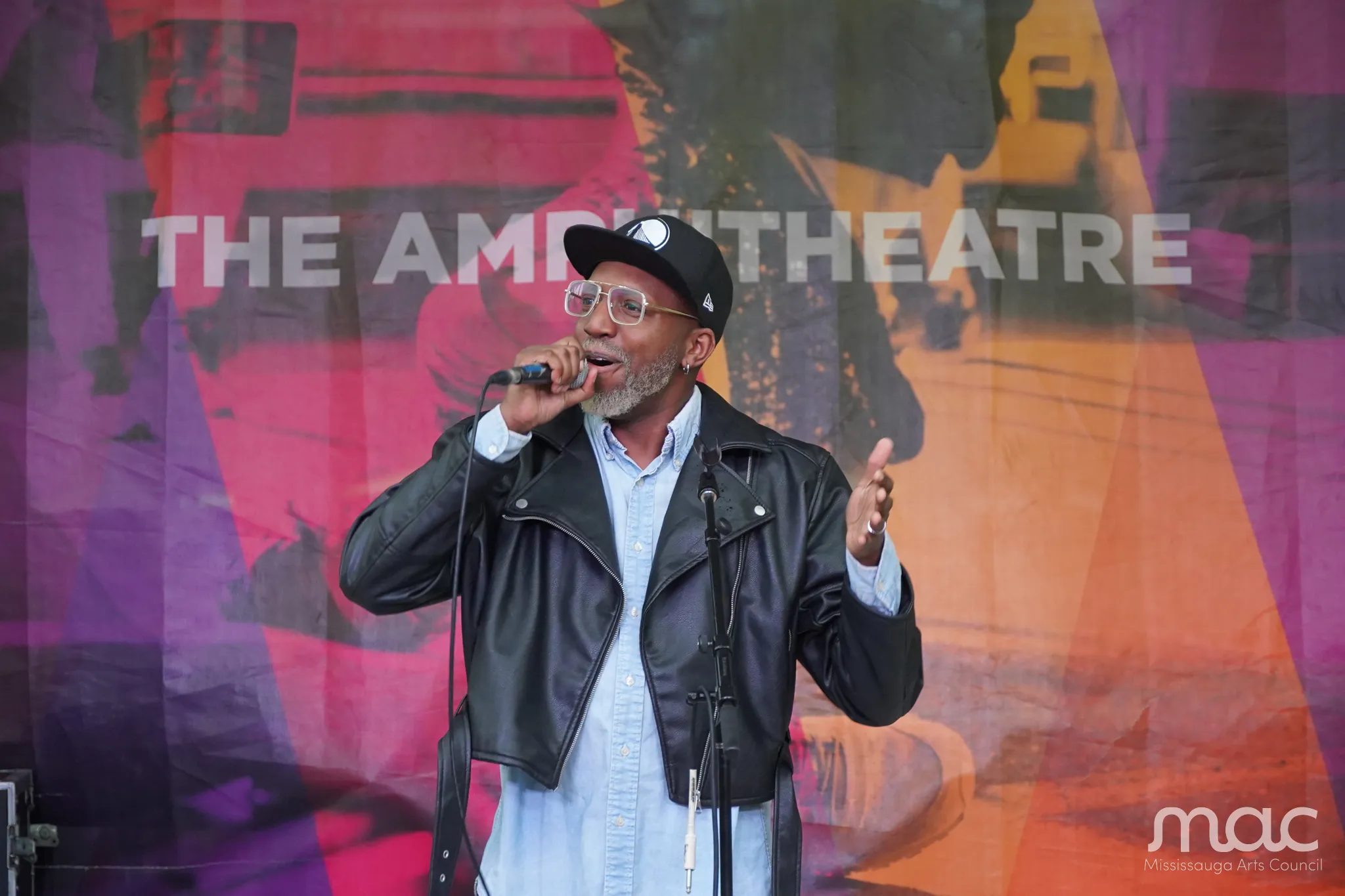 A performer is singing into a microphone. They wear a black snap back hat, a black leather jacket, and a denim shirt. Behind them, an outdoor banner reads "The Amphitheatre" A performer is singing into a microphone. They wear a black snap back hat, a black leather jacket, and a denim shirt. Behind them, an outdoor banner reads "The Amphitheatre"