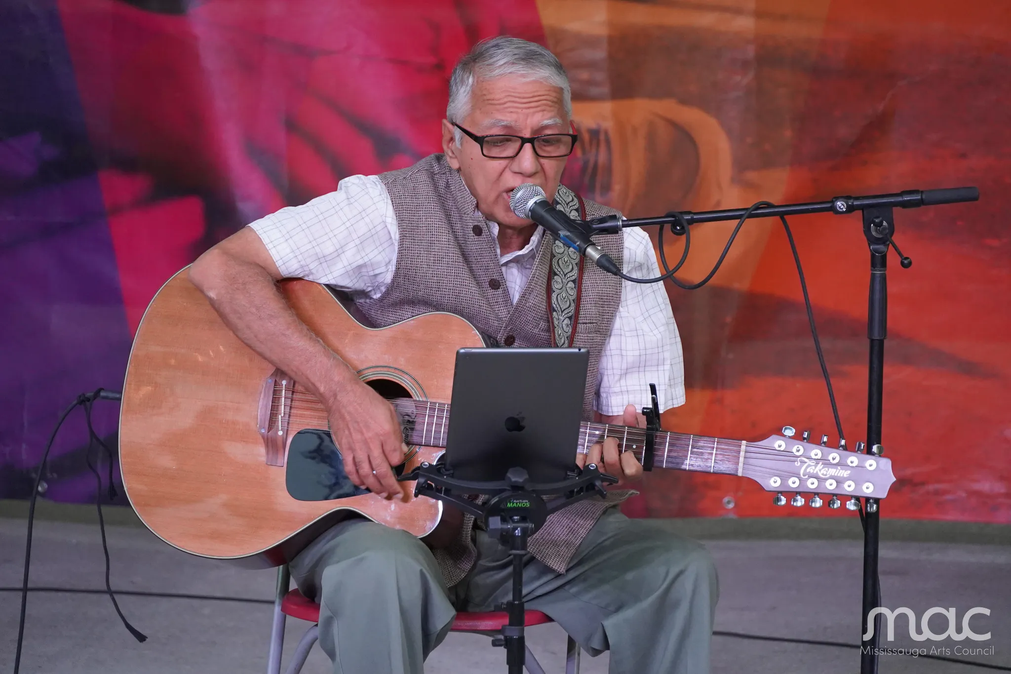 An older man with glasses is sitting on a chair and playing an acoustic guitar while singing into a microphone.