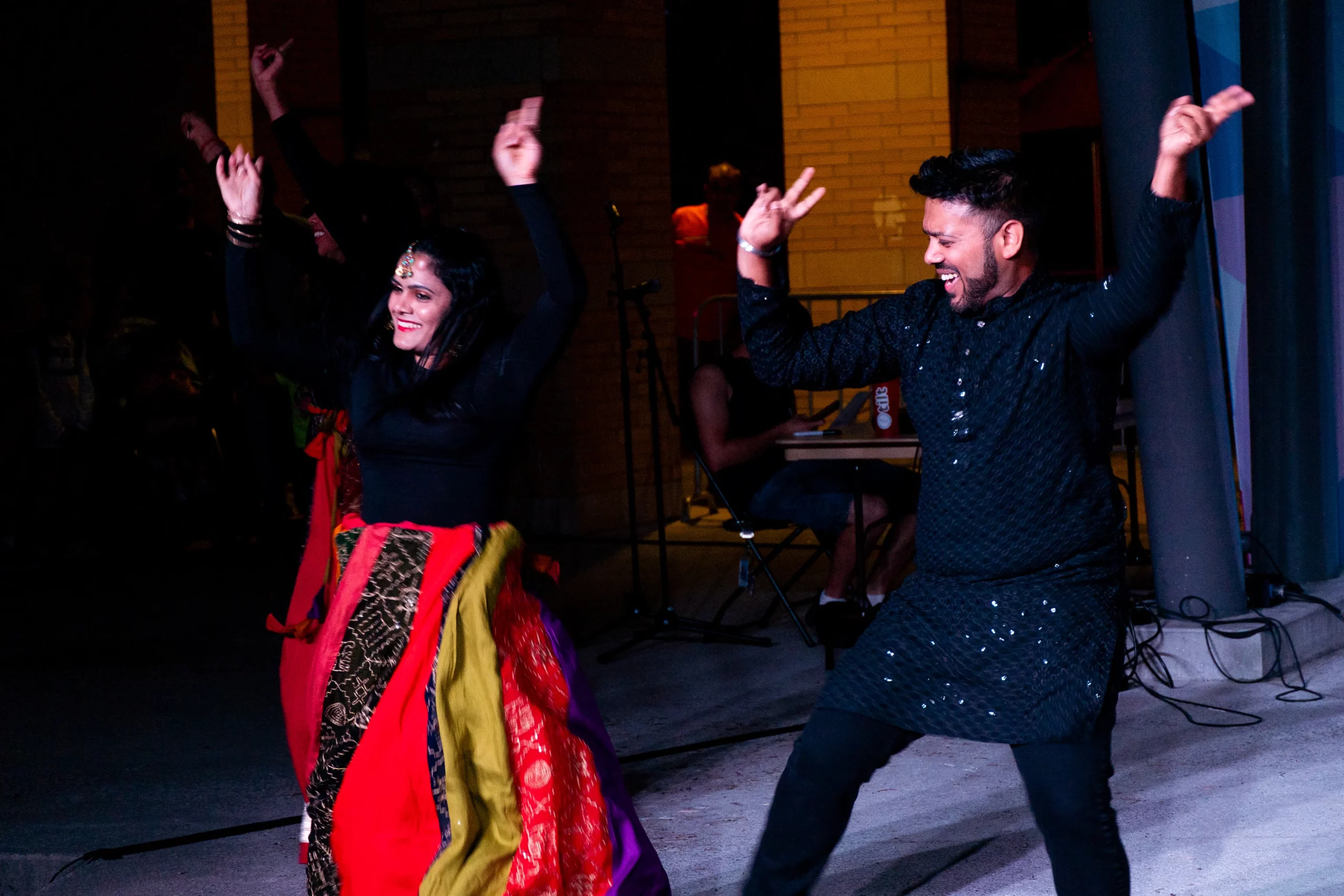Two dancers in traditional South Asian attire smile and dance side by side with their arms raised. The performance is in an amphitheatre at night. Two dancers in traditional South Asian attire smile and dance side by side with their arms raised. The performance is in an amphitheatre at night.