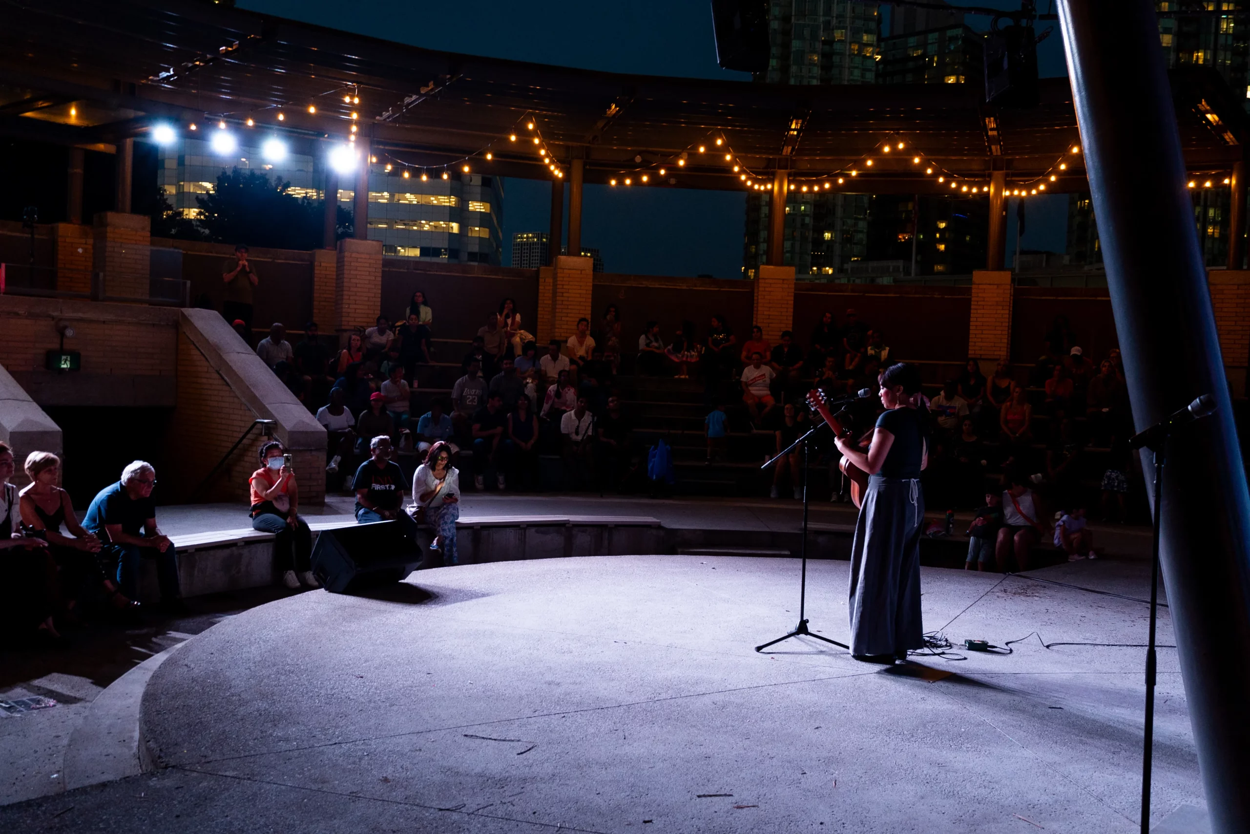 A young woman performers with her guitar in an outdoor open mic at night. She is lit from behind, with a diverse crowd seated in front of her. A young woman performers with her guitar in an outdoor open mic at night. She is lit from behind, with a diverse crowd seated in front of her.