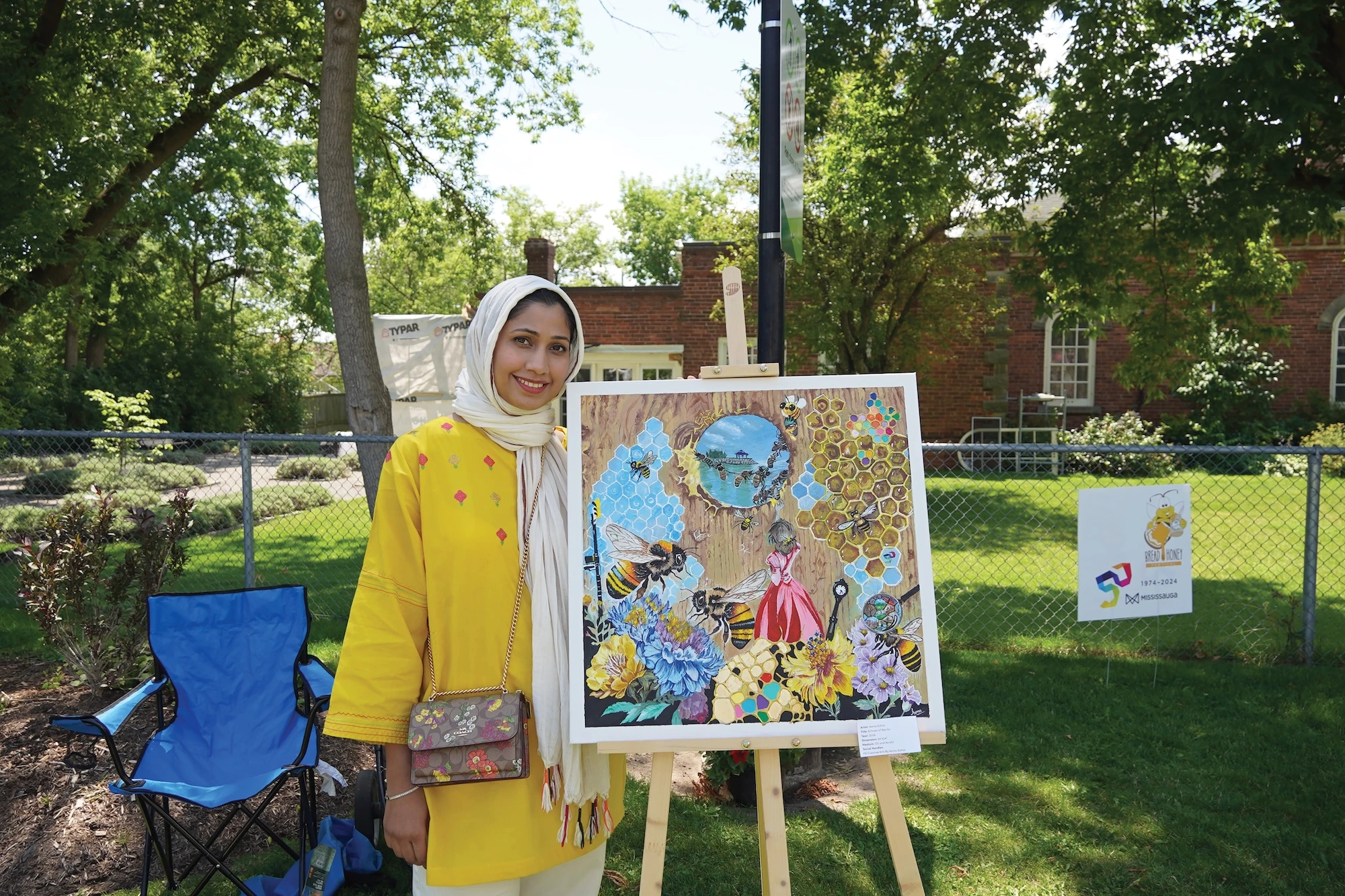 A woman in a yellow tunic and white hijab stands next to her colorful painting of bees, honeycombs, and flowers displayed on an easel outdoors.